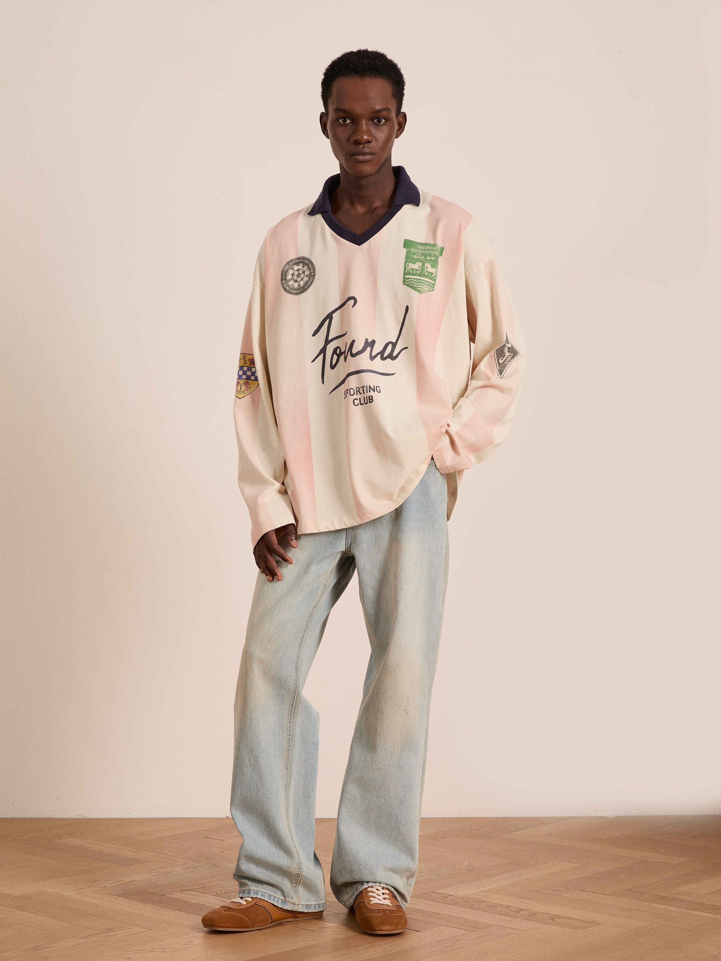 A person poses indoors against a plain wall and wood floor wearing the FOUND Striped Sport LS Jersey in beige and pink, paired with light blue jeans and brown shoes for a unisex relaxed fit look.