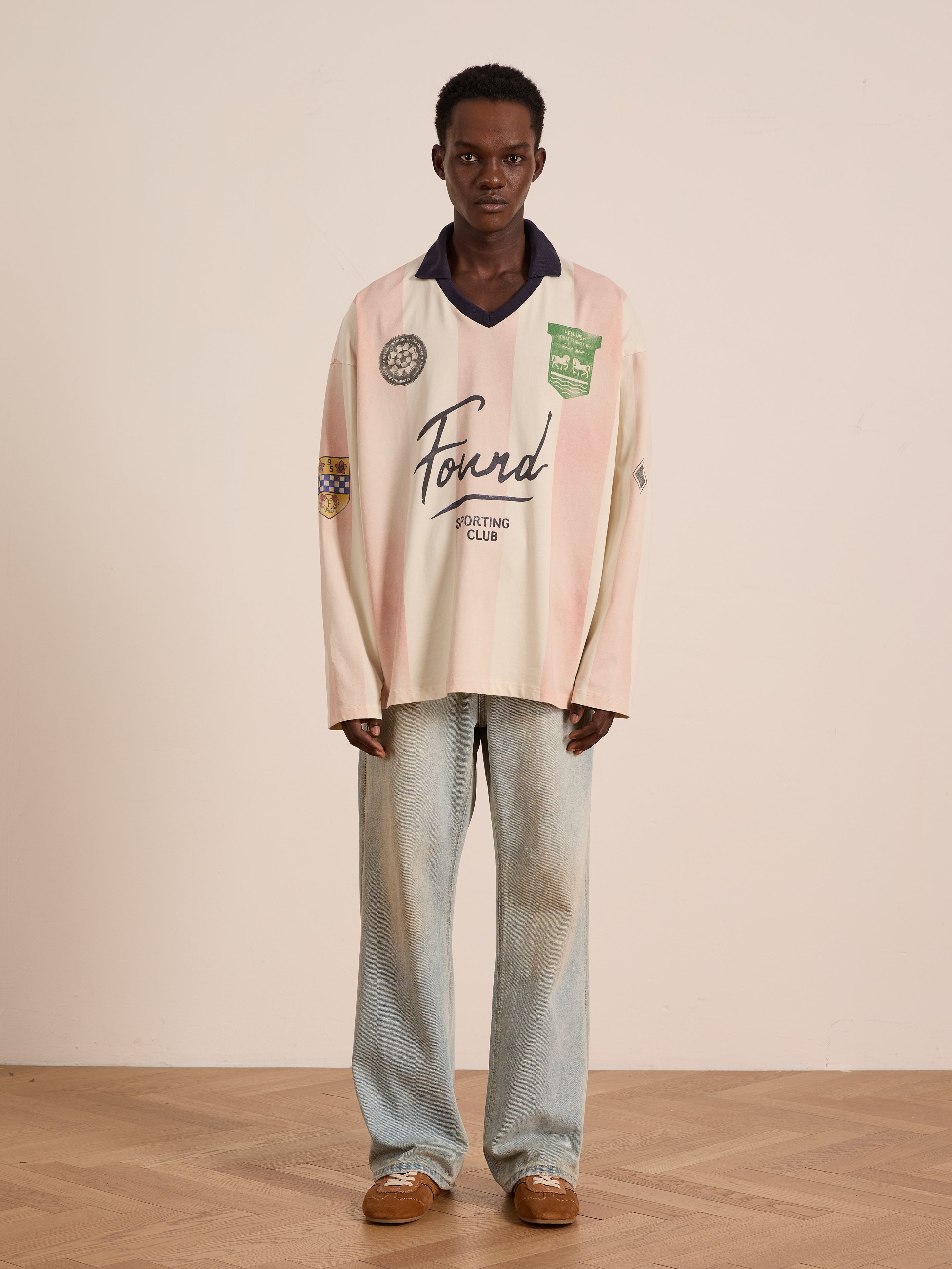 A person stands indoors in the FOUND Striped Sport LS Jersey, featuring a relaxed fit and football silhouette graphics, paired with light blue jeans and brown shoes on a wood floor against a plain wall.