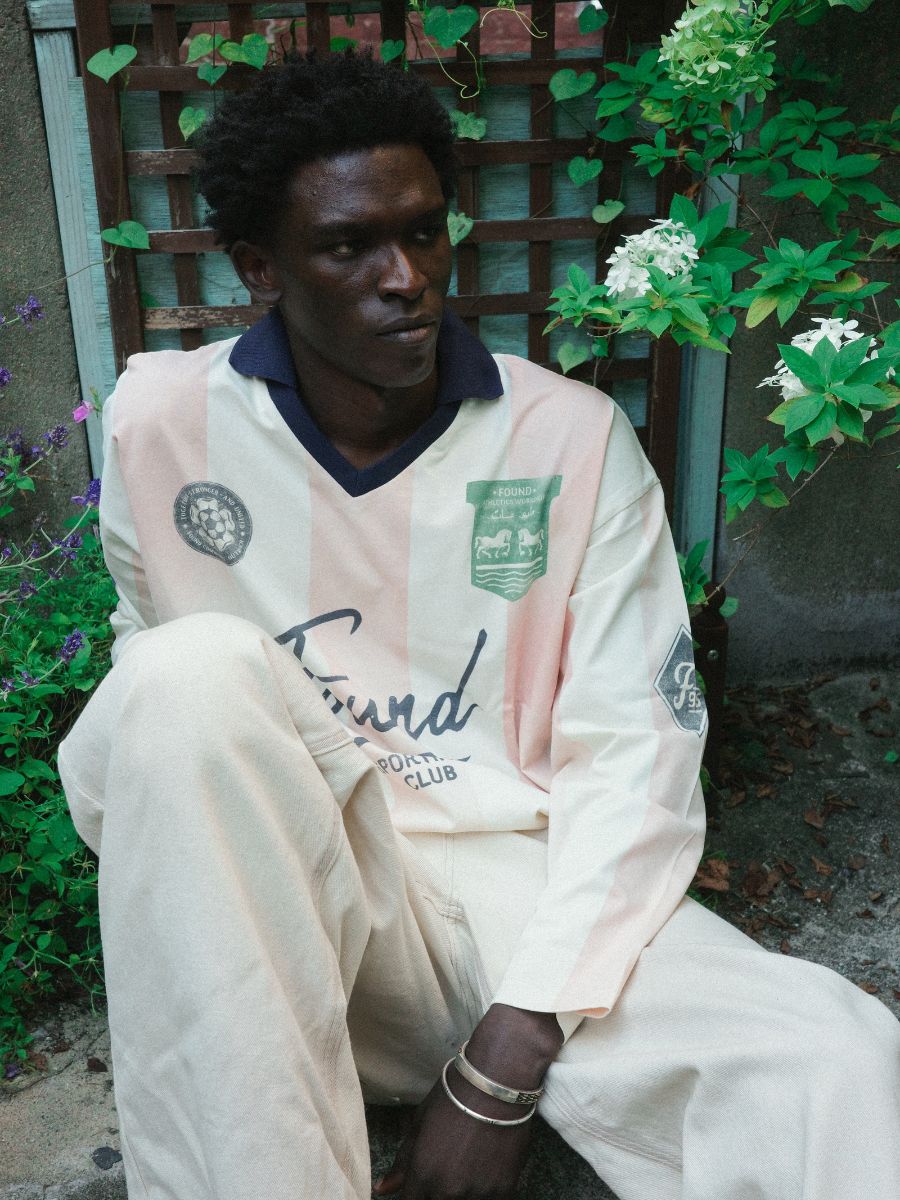 A man wears the FOUND Striped Sport LS Jersey, a unisex relaxed-fit long-sleeve rugby shirt in cream and pale pink with logos, while sitting outdoors by a wooden trellis and green flowering plants.