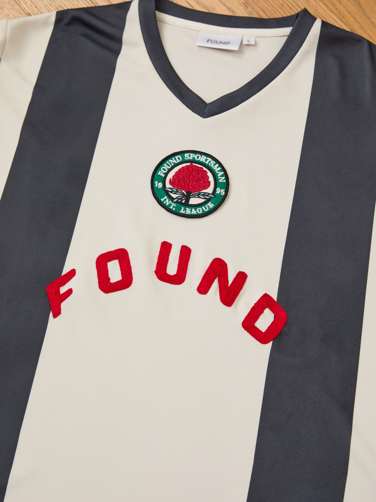 The Striped Stateman Jersey by FOUND features beige and black stripes, a red “FOUND” logo, and a green “FOUND SPORTSMAN AM. LEAGUE 1995.” patch.
