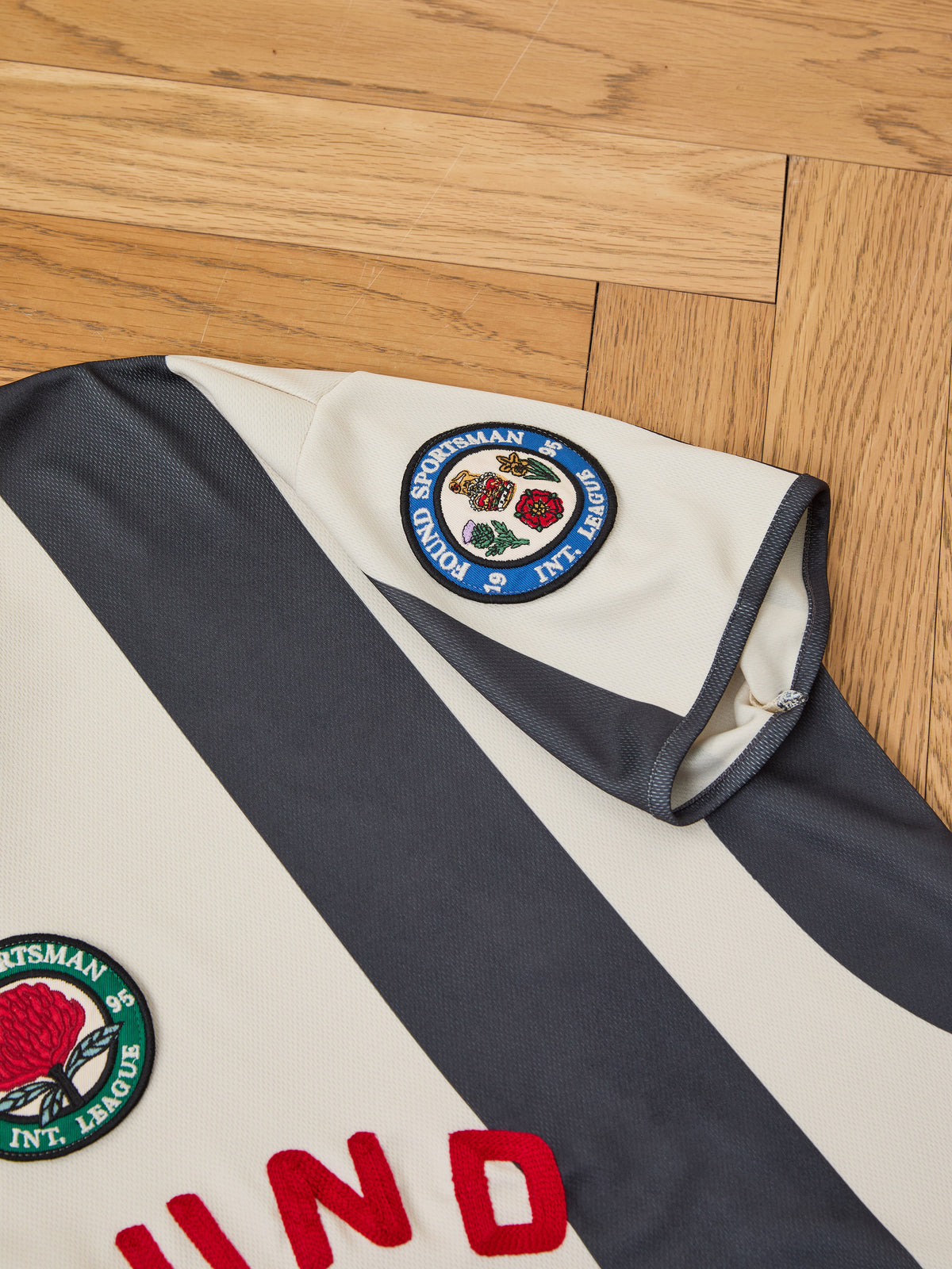 Close-up of the Striped Stateman Jersey by FOUND—cream and dark gray stripes, vintage unisex sportswear with embroidered patches, displayed on a wooden floor.