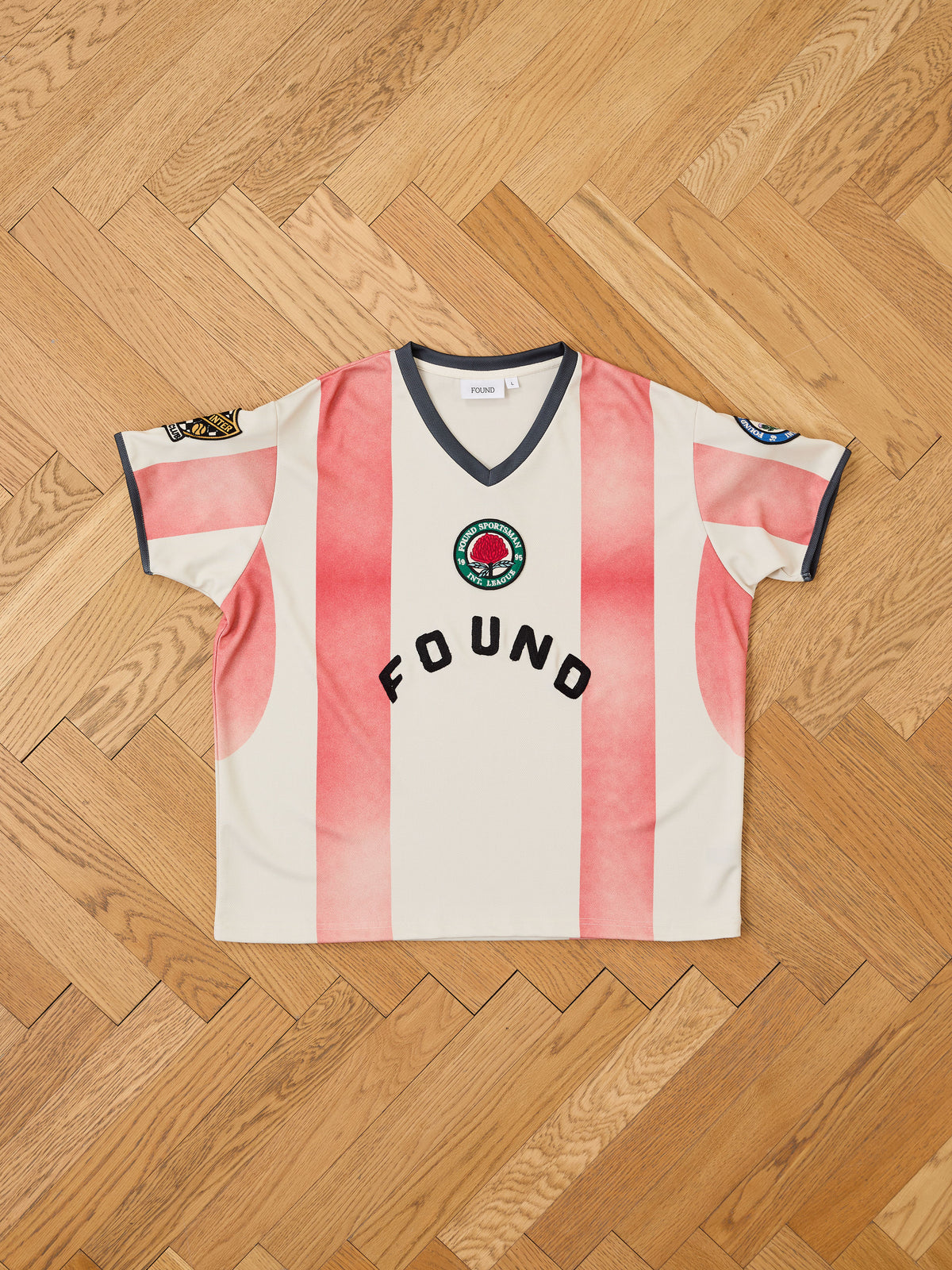 A short-sleeve FOUND Striped Stateman Jersey, featuring wide pink and white stripes with black trim and patches, is laid flat on a wooden herringbone floor. Inspired by vintage football jerseys, this unisex sportswear piece displays “FOUND” printed prominently across the chest.
