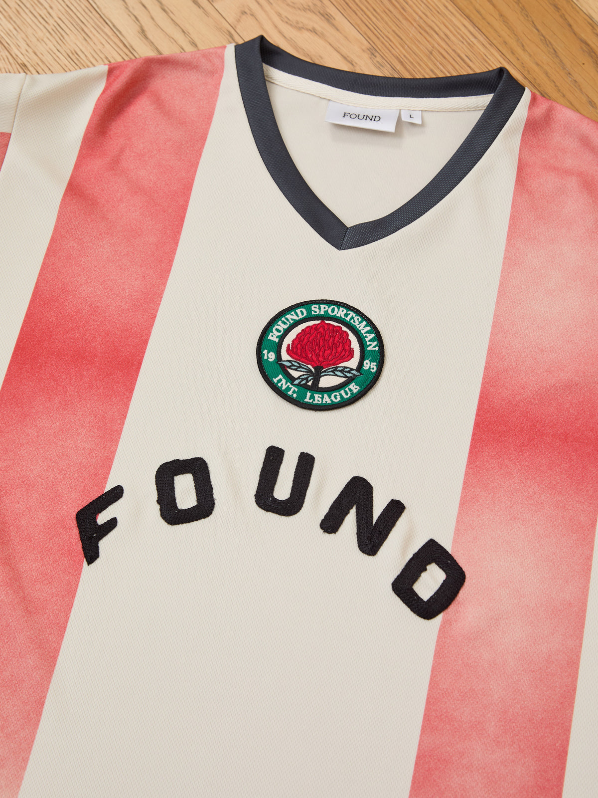 The FOUND Striped Stateman Jersey, a cream and red striped sports jersey with a black V-neck, showcases the “FOUND” text and a round patch reading “FOUND SPORTSMAN ATH. LEAGUE” with a striking red emblem. Laid on a wooden surface, this exceptional unisex piece captures the vintage spirit of classic football jerseys.