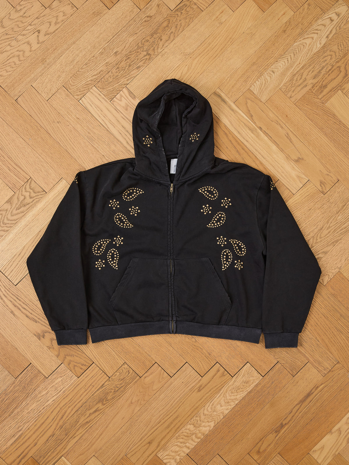 The FOUND Studded Zip Up Hoodie with gold paisley accents is shown flat on a wooden herringbone floor for a vintage-inspired look.