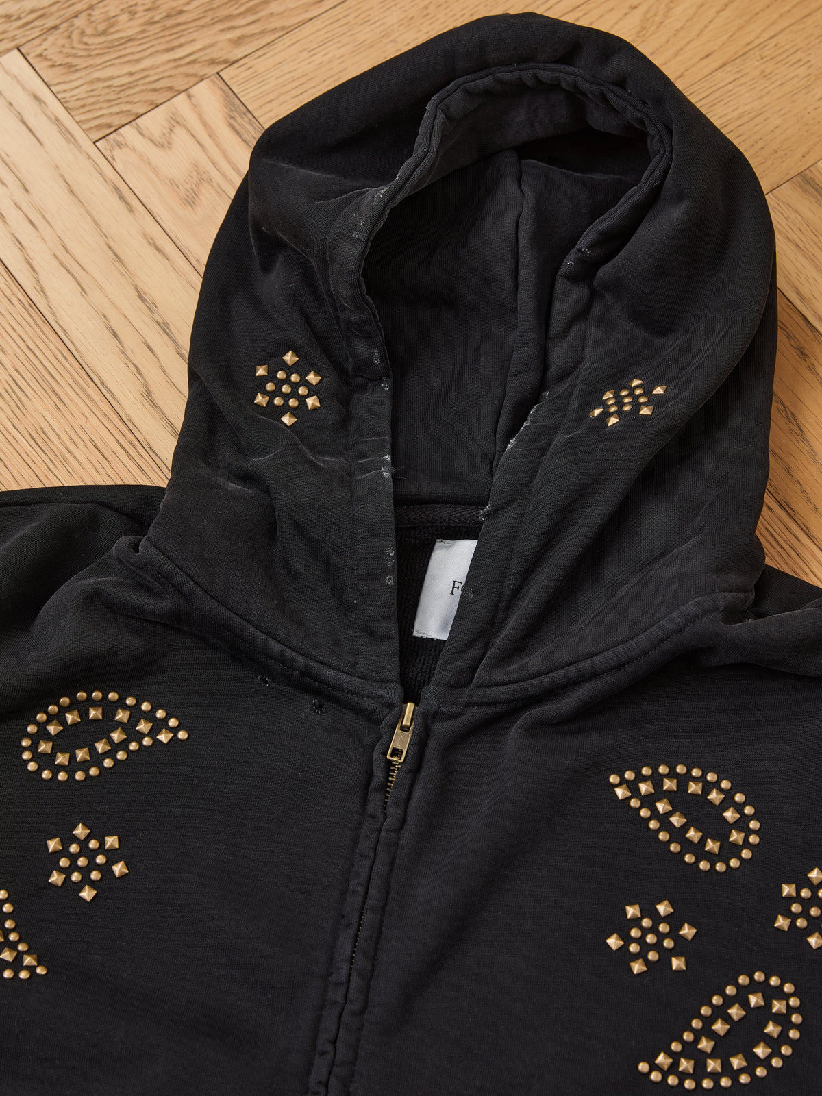 FOUND Studded Zip Up Hoodie in black with gold square stud patterns, shown laid flat on wood floor. Hood is up, partially covering a white collar tag.