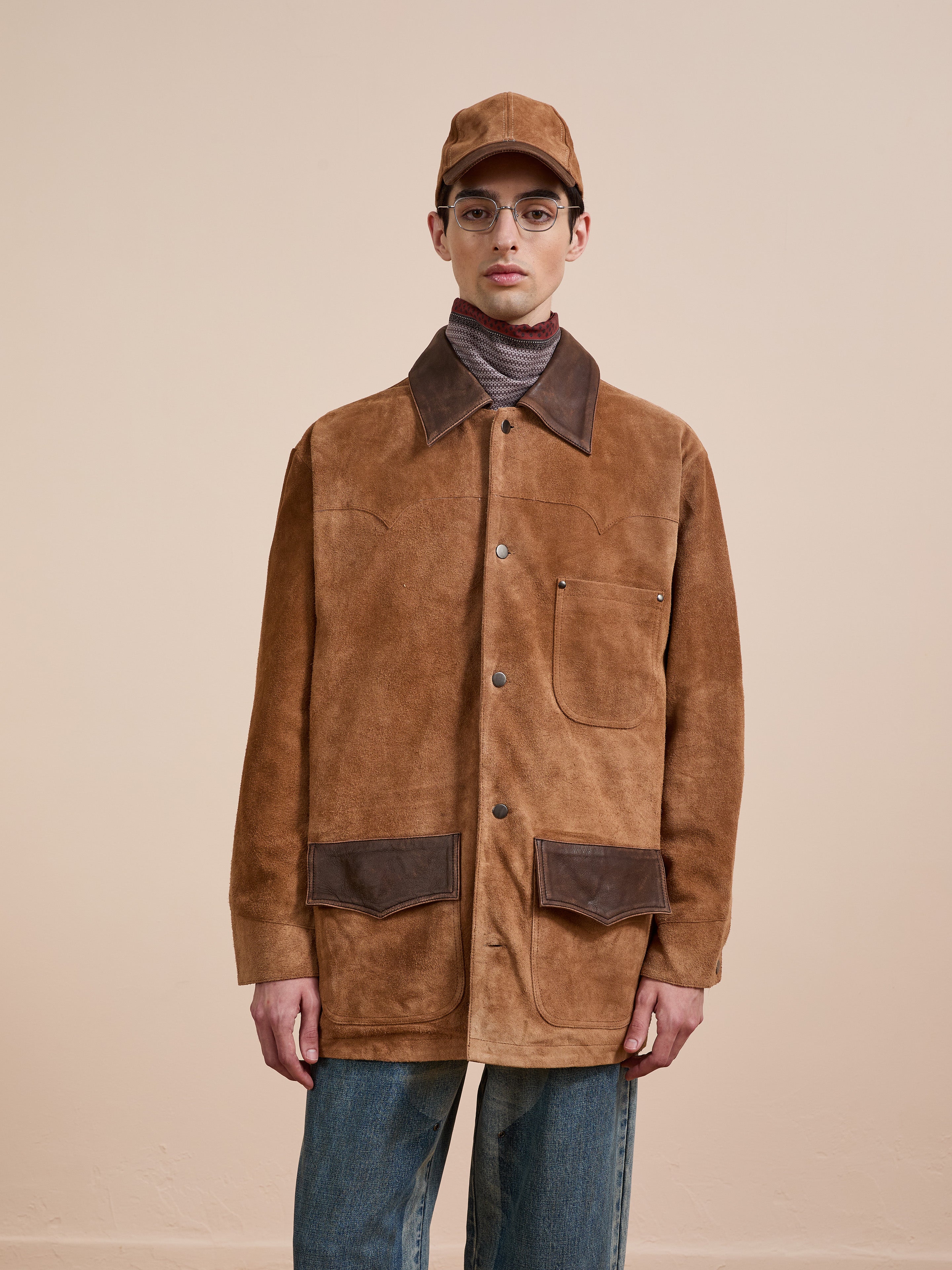 Someone in the FOUND Suede Contrast Chore Jacket, featuring dark brown accents, pairs it with blue jeans, a brown cap, and glasses while standing against a plain beige background.