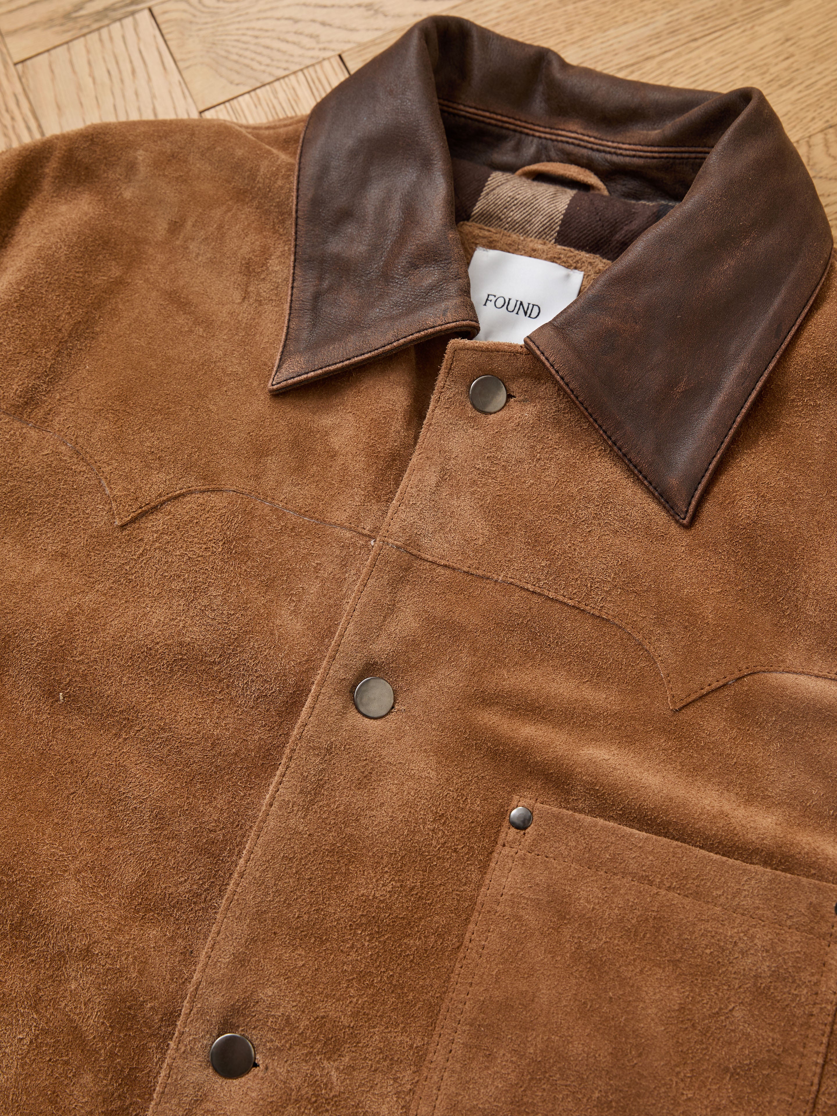 The FOUND Suede Contrast Chore Jacket features a vintage-inspired design with a dark brown leather collar, metal snap buttons, and chest pocket, shown laid flat on a wooden surface.