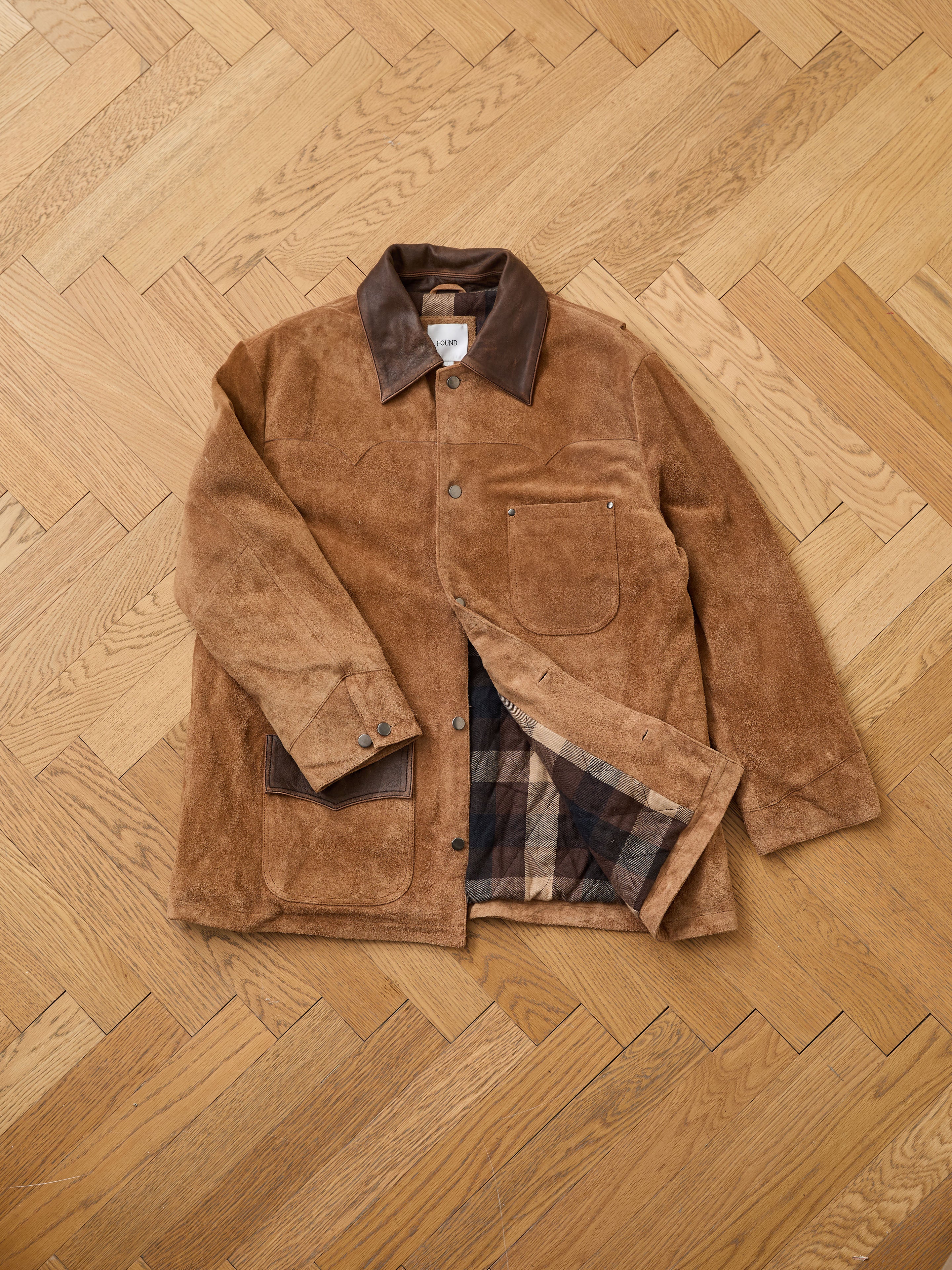 The FOUND Suede Contrast Chore Jacket is vintage-inspired, with brown suede, a dark leather collar and cuffs, front pocket, and plaid lining—shown laid flat on a wooden herringbone floor.