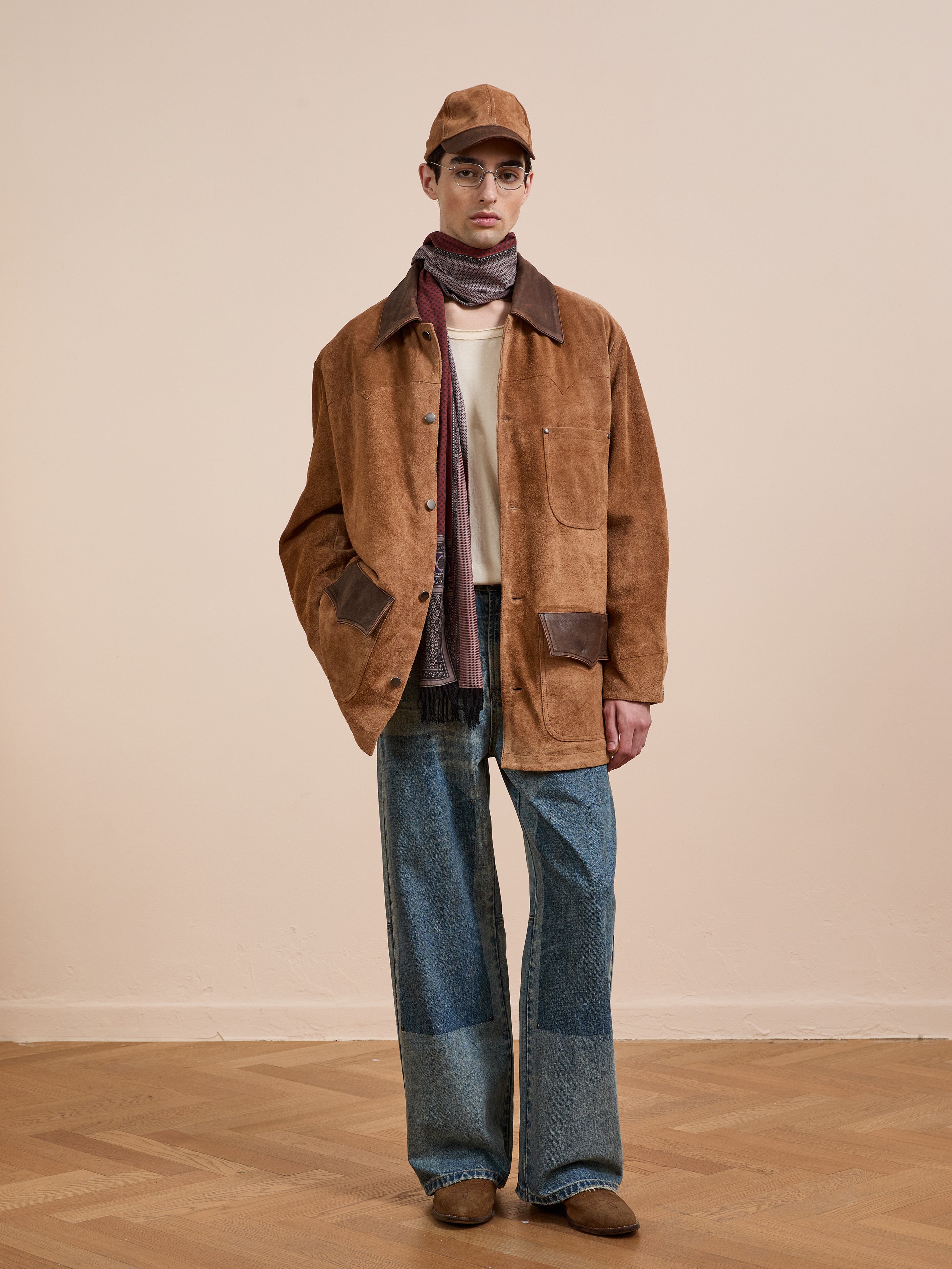 Someone wearing the FOUND Suede Contrast Chore Jacket, wide-leg patchwork jeans, brown shoes, a tan cap, and a scarf stands on a wooden floor against a beige wall.