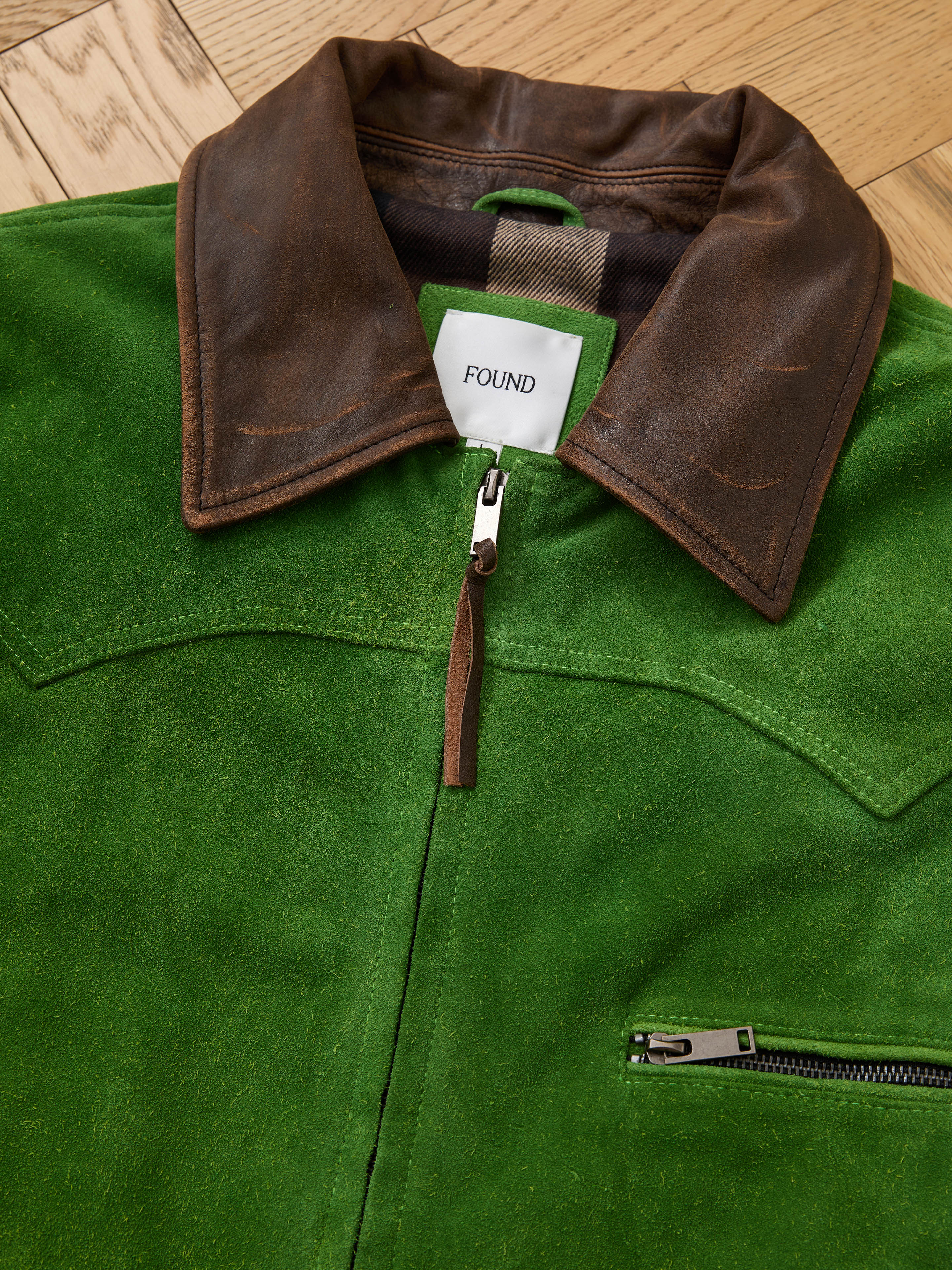 The FOUND Suede Western Barn Jacket features green suede leather, a brown leather collar, western yoke stitching, a chest zipper pocket, and an interior "FOUND" label, shown laid flat on a wooden floor.