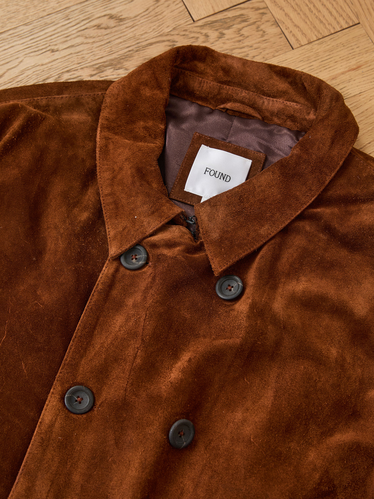 The Belted Suede Trench Coat by FOUND is a brown suede double-breasted unisex style with black buttons, shown on a wooden floor.