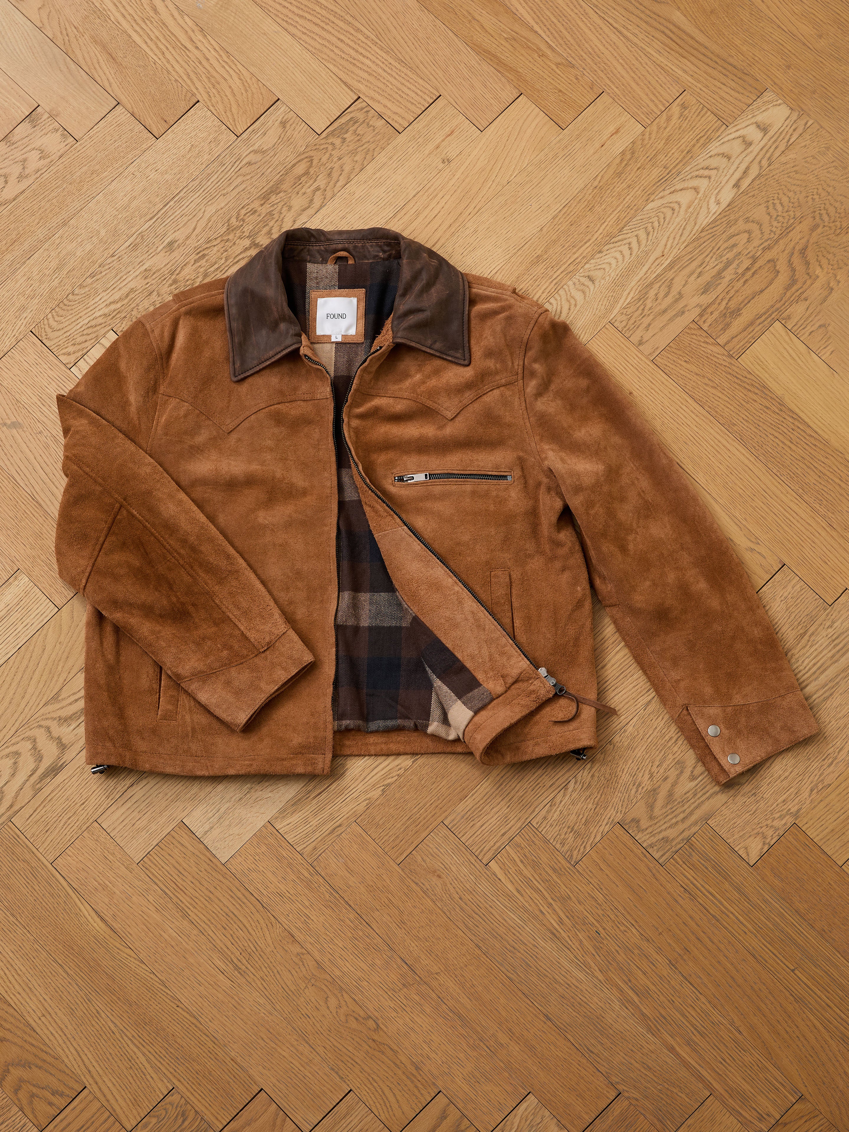 The FOUND Suede Western Barn Jacket, a unisex relaxed fit in brown suede with a dark collar, plaid lining, zip chest pocket, and snap cuffs, is shown on a wooden herringbone floor—offering comfort and timeless appeal.