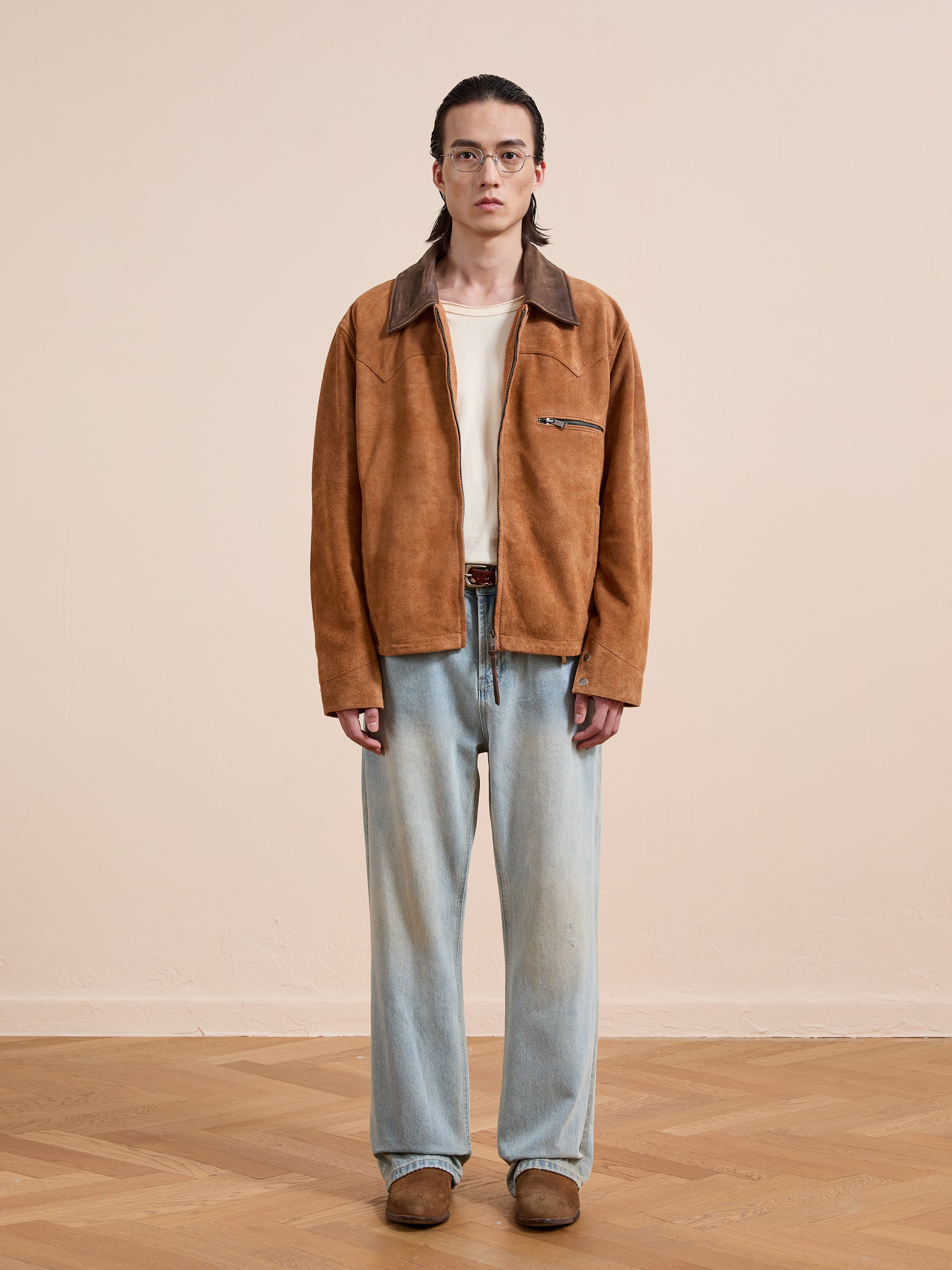 A person stands indoors against a plain beige background, wearing glasses, a FOUND Suede Western Barn Jacket over a white shirt, light blue jeans, and brown shoes.