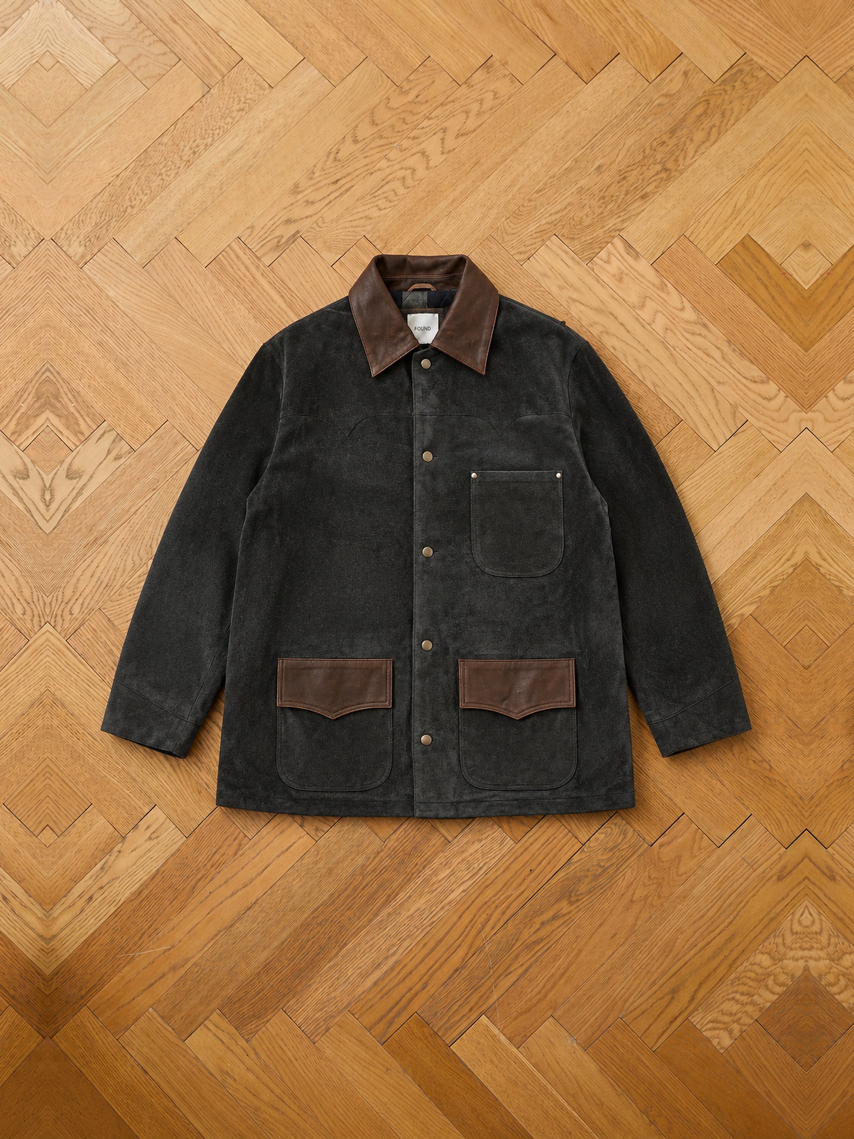 The FOUND Suede Contrast Barn Jacket, featuring a dark corduroy body with brown leather collar and pocket accents, reminiscent of a vintage barn jacket, is displayed flat on a wooden herringbone floor.