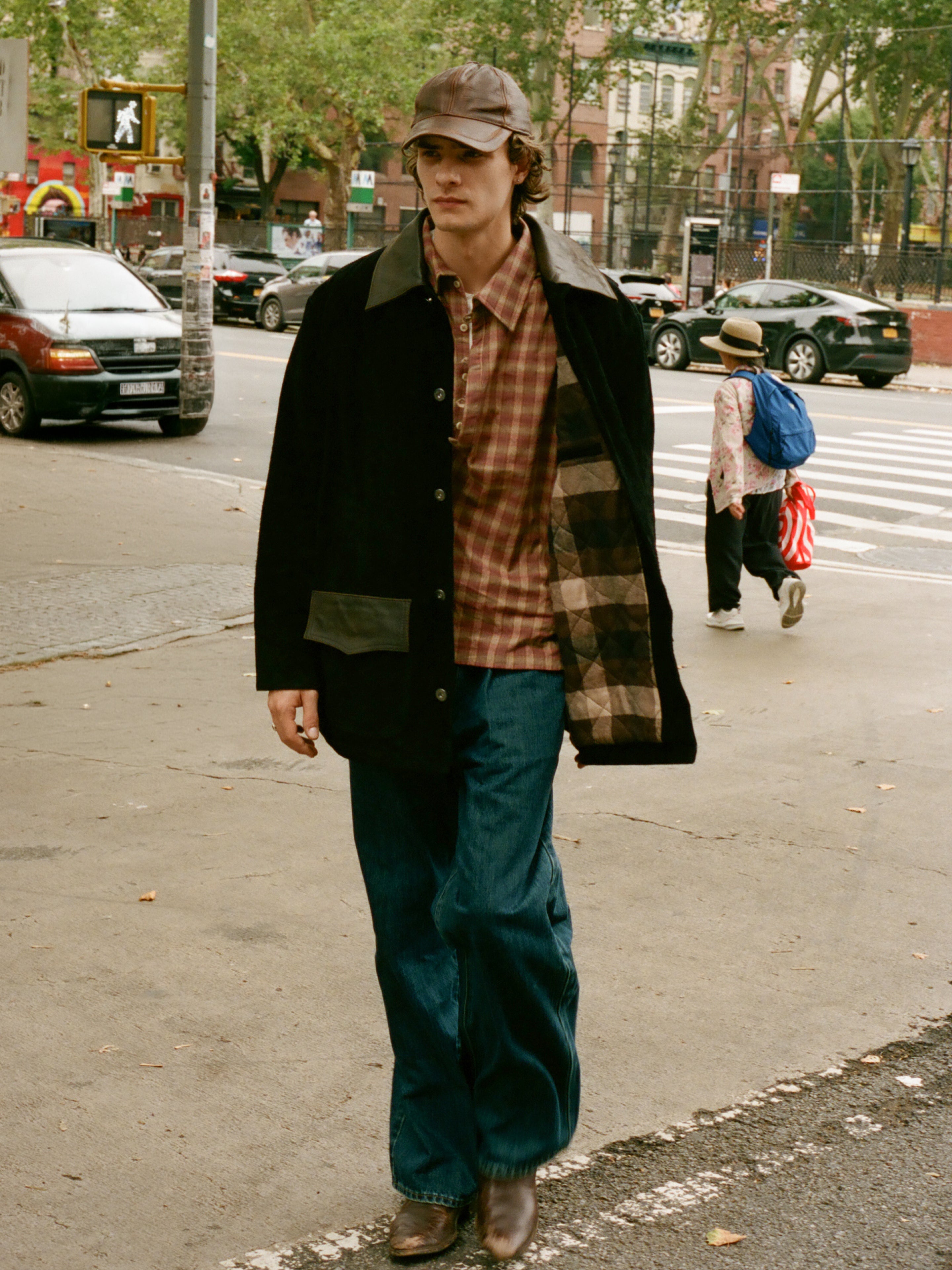 Wearing the FOUND Suede Contrast Barn Jacket, a person in a cap, plaid shirt, and wide blue jeans walks along a city sidewalk near a crosswalk.