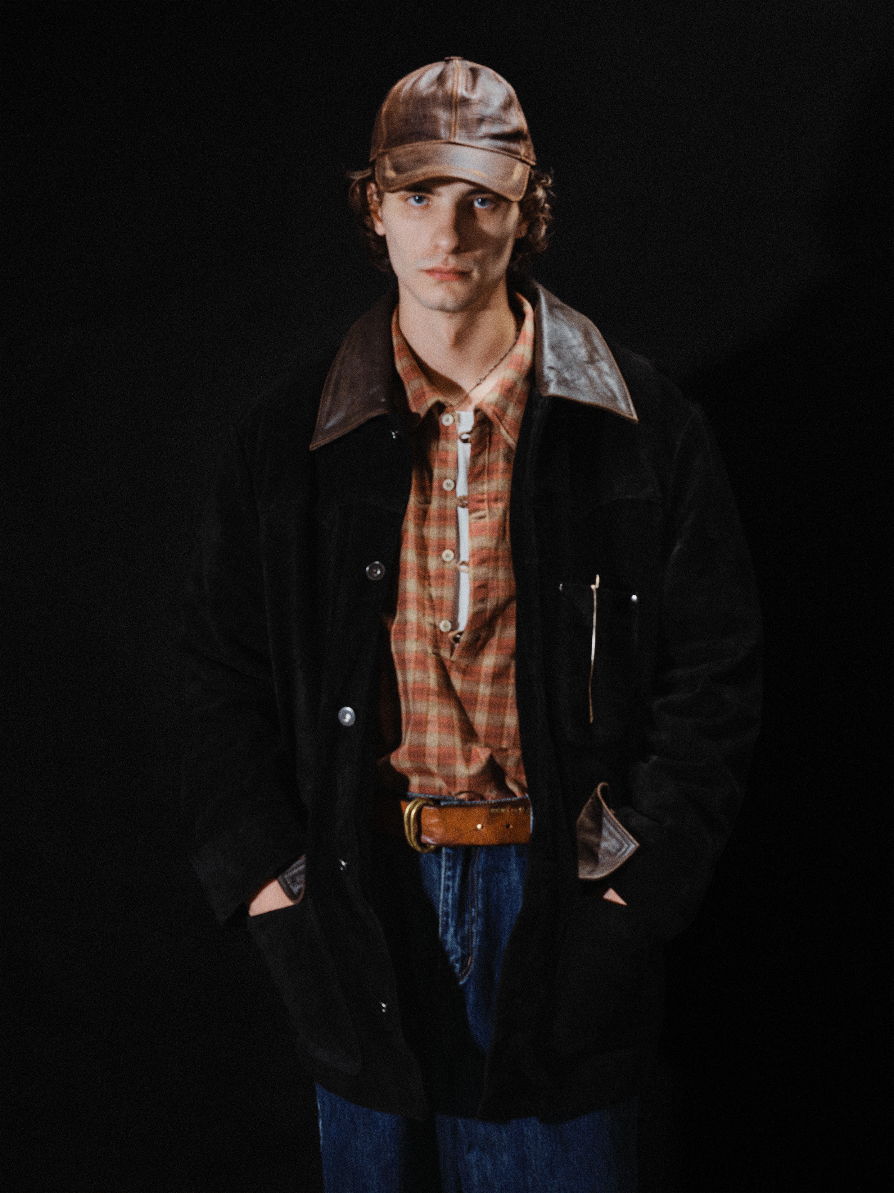 A person wearing a leather cap, plaid shirt, classic brown belt, blue jeans, and the FOUND Suede Contrast Barn Jacket stands with hands in coat pockets against a black background.