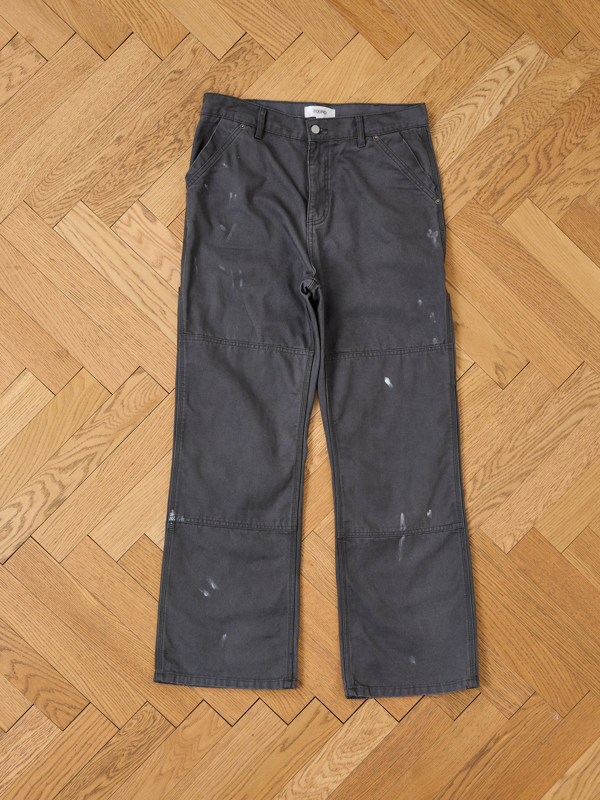 A pair of FOUND Sunbleached Utility Pants in dark gray with paint splatters lies flat on a wooden herringbone floor.