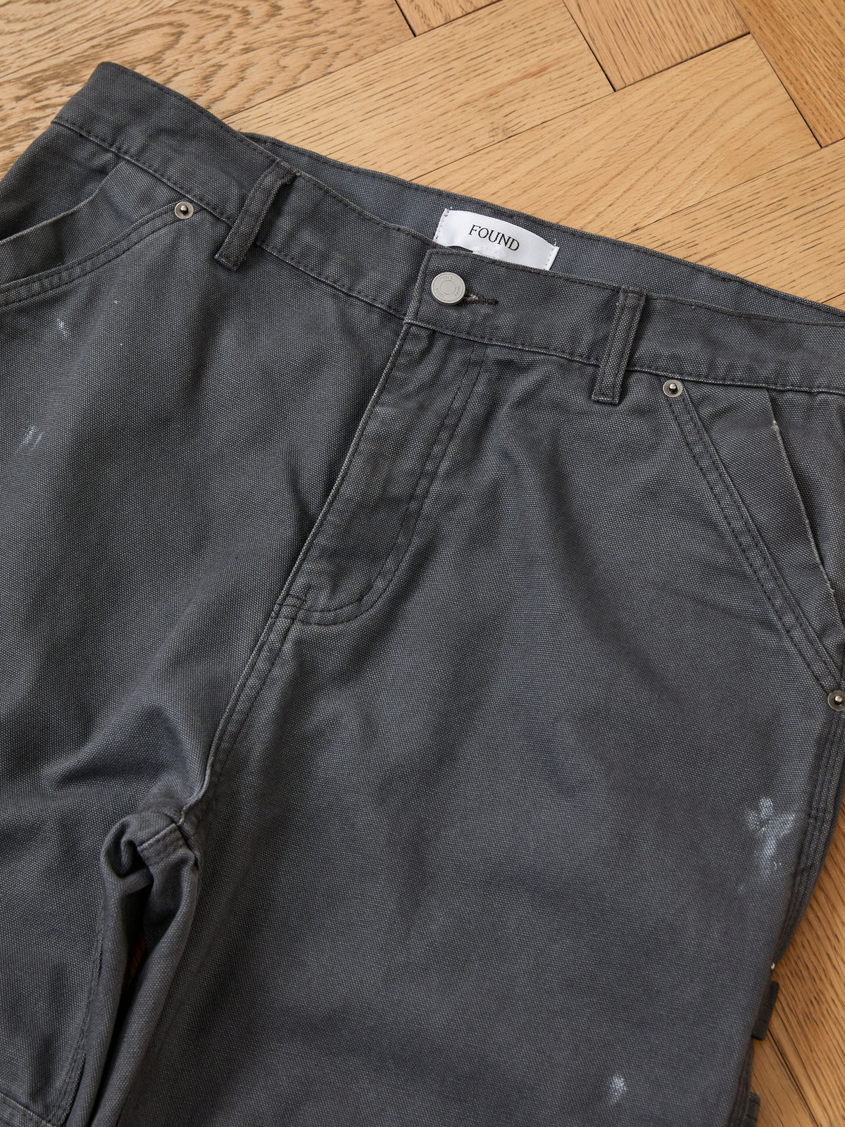 A pair of Sunbleached Utility Pants by FOUND is laid flat on a wooden floor, displaying pockets, belt loops, and metal hardware.