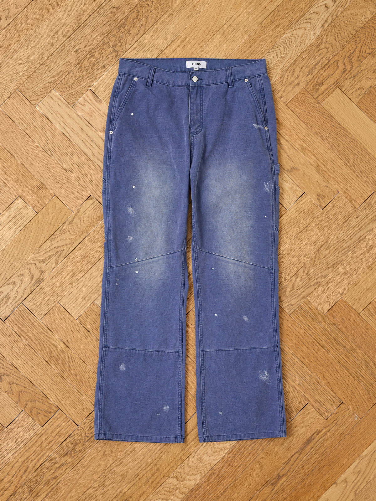 FOUND Sunbleached Utility Pants feature blue painter-style utility design with white paint splatters and faded spots, laid flat on a herringbone-patterned wooden floor.