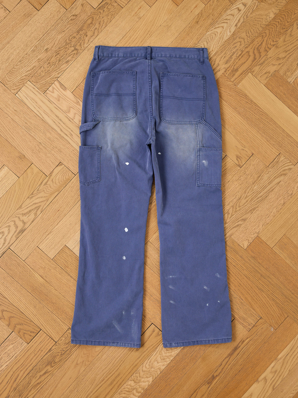 FOUND Sunbleached Utility Pants, relaxed fit in blue with paint stains and faded details, shown laid flat on a wooden herringbone floor.