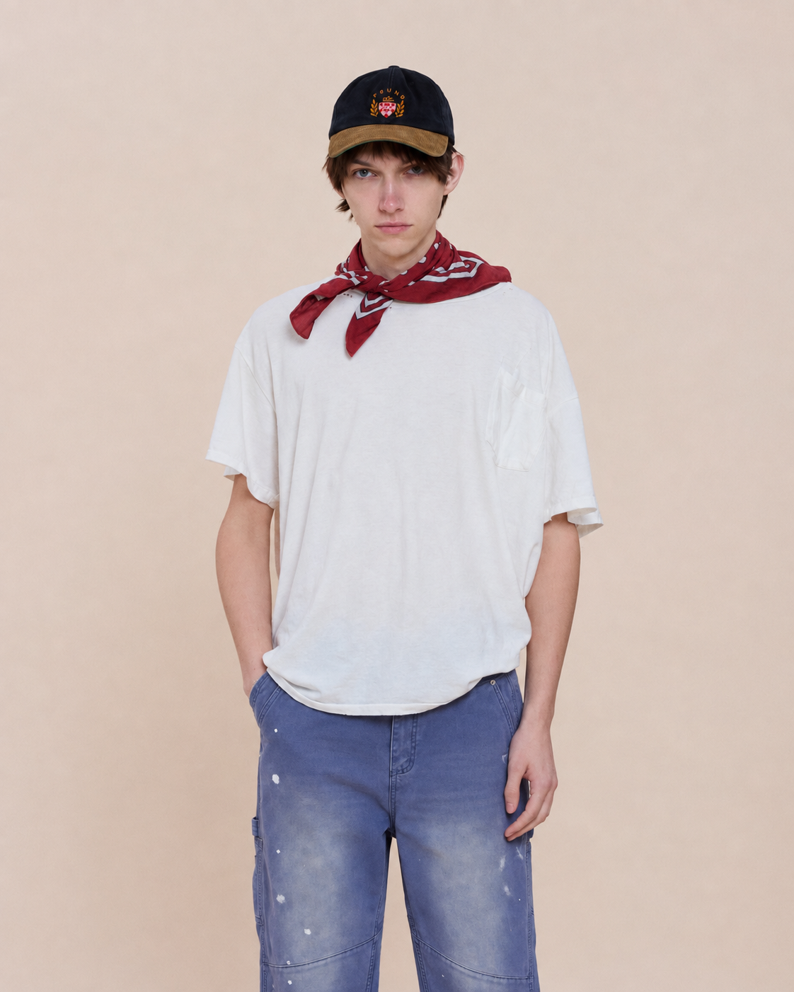 Young person in a black cap, red bandana, white t-shirt, and FOUND Sunbleached Utility Pants stands against a plain beige background with hands in pockets.