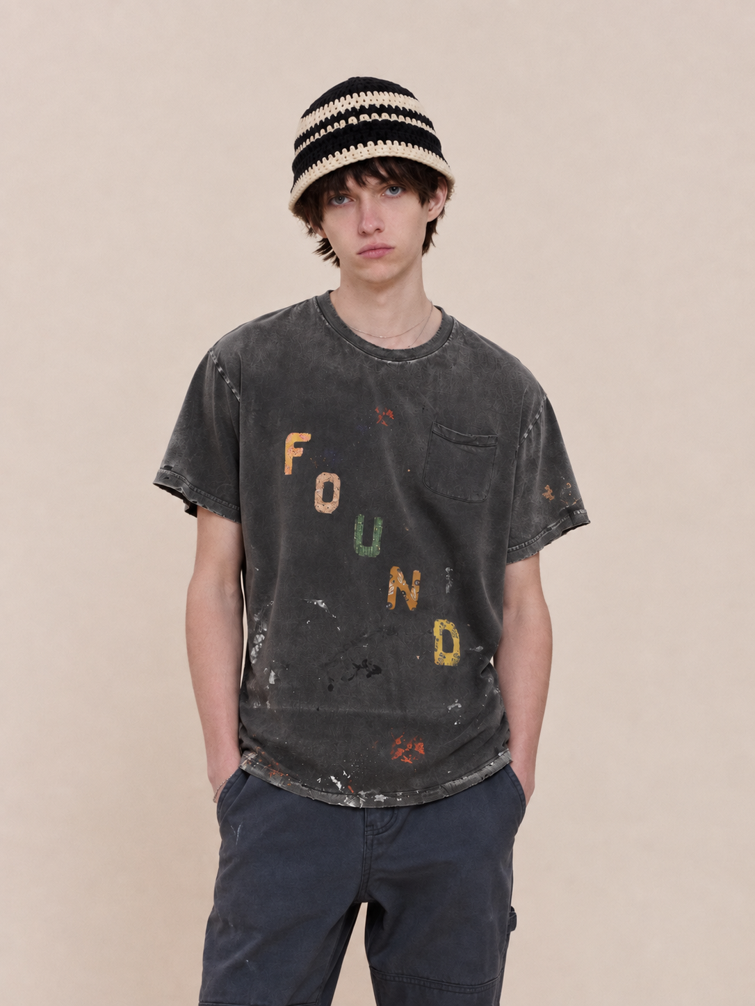 Young person in a striped bucket hat and faded black Tapestry Logo Paint Tee by FOUND, unisex relaxed fit, stands against a plain beige background.