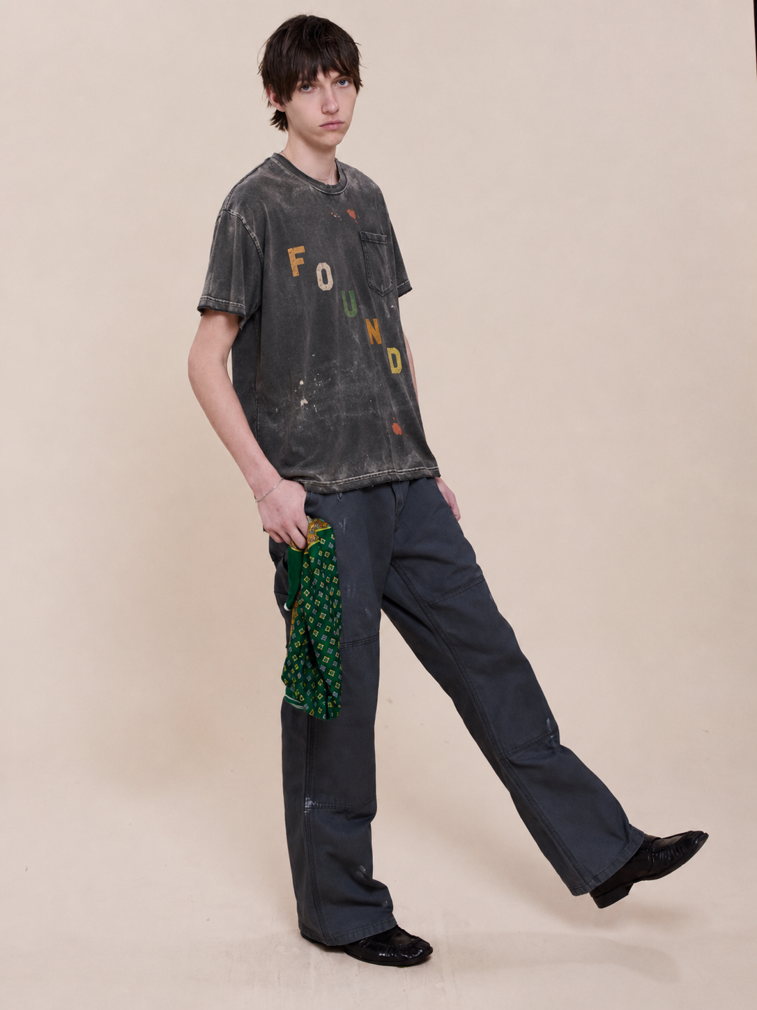 Person wears the FOUND Tapestry Logo Paint Tee, unisex relaxed fit with distressed paint splatter, styled with dark pants against a beige background, holding a green patterned cloth.