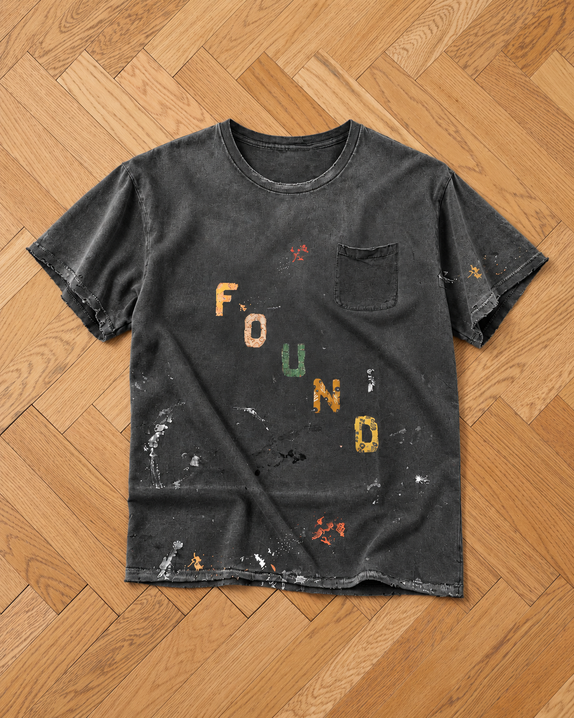 The Tapestry Logo Paint Tee by FOUND: black, paint-splattered, chest pocket, “FOUND” in colorful letters, unisex relaxed fit on herringbone wood floor.
