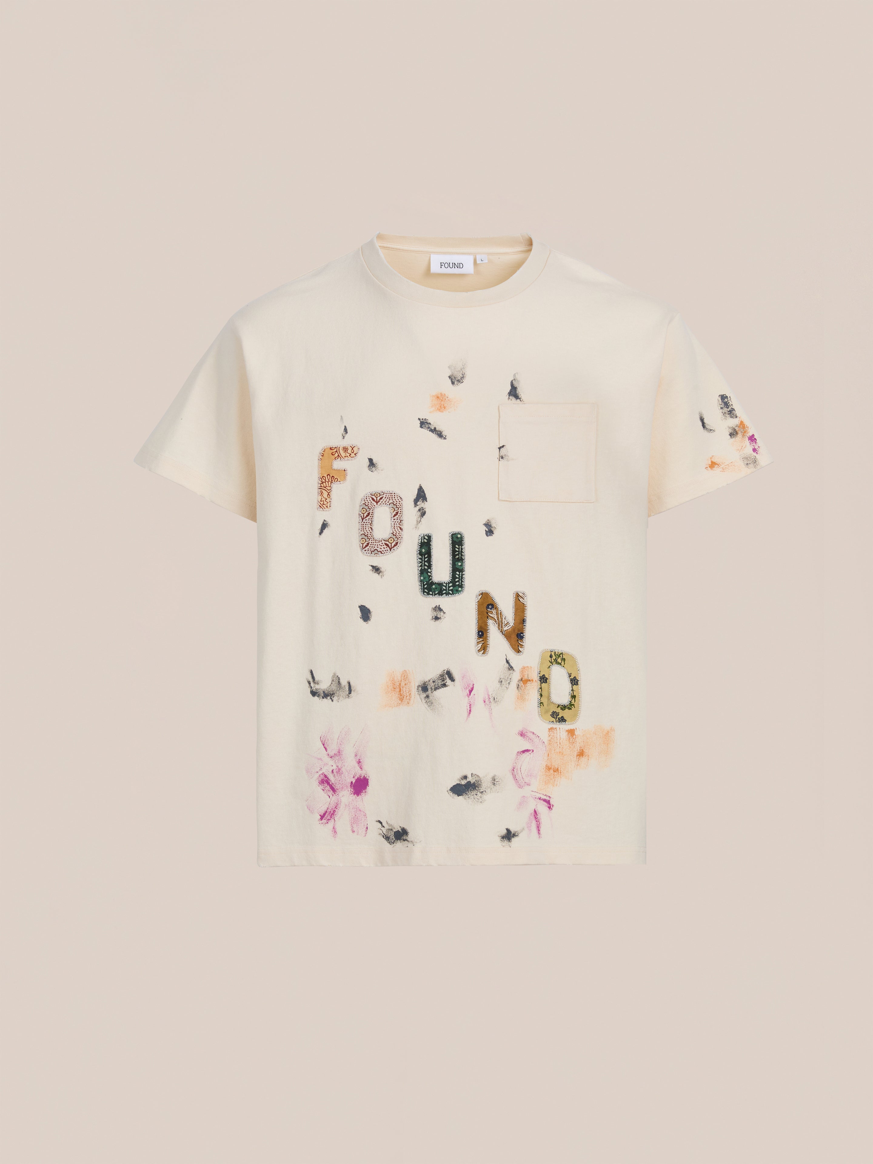 The FOUND Tapestry Logo Paint Tee is cream-colored, made from premium cotton, with a relaxed fit, chest pocket, and "FOUND" logo in mixed patterns plus floral and abstract prints.