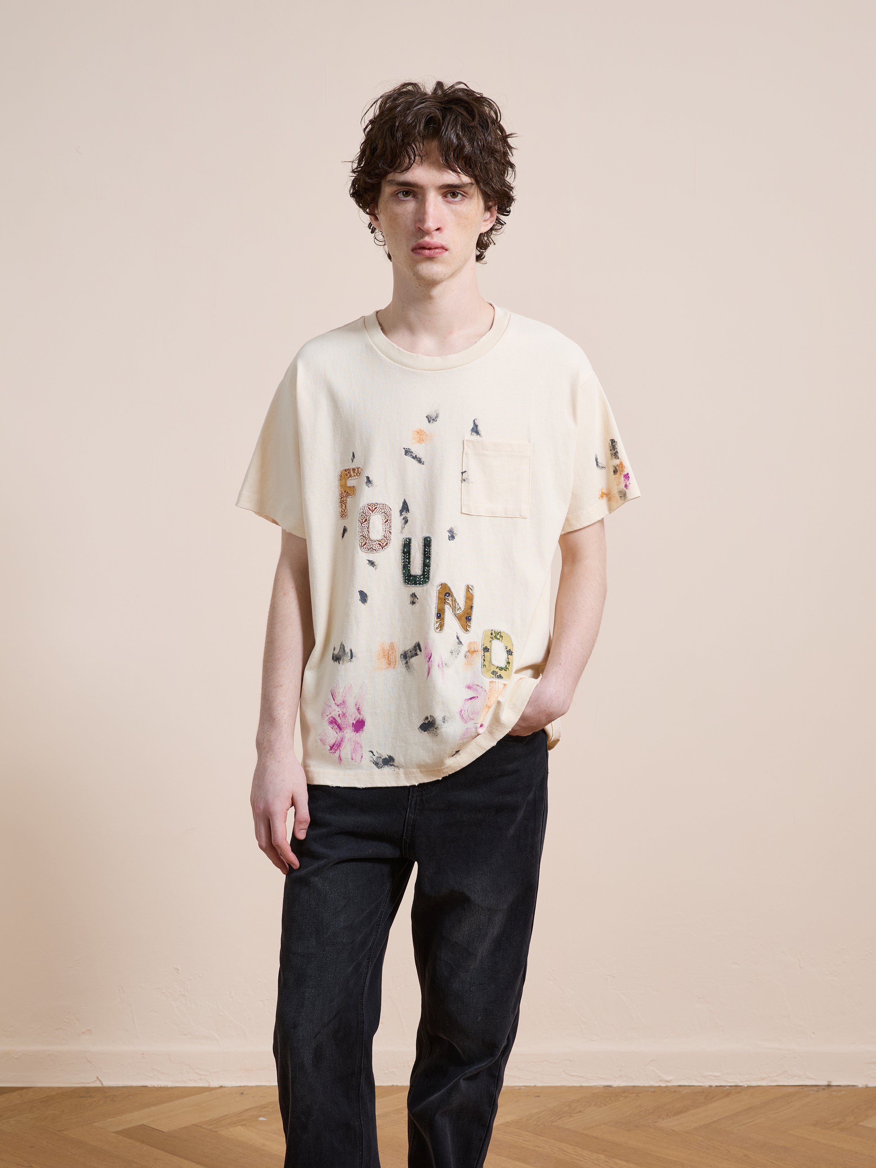 A young person stands indoors wearing the FOUND Tapestry Logo Paint Tee, a cream premium cotton shirt with colorful "FOUND" lettering and black jeans, highlighting its unisex relaxed fit with one hand in their pocket.