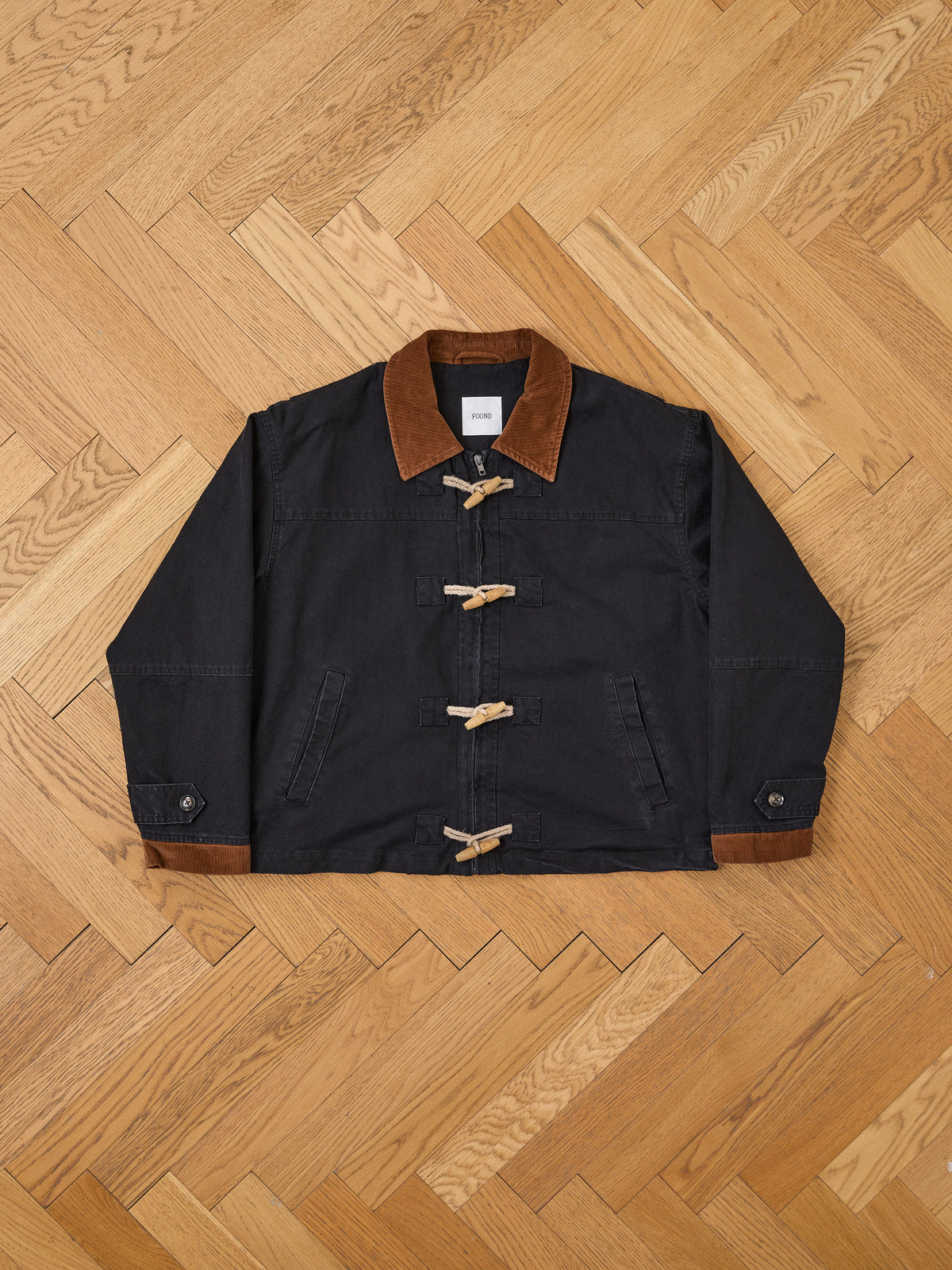 The Found Toggle Work Jacket, featuring a black vintage design, brown corduroy collar, and fireman-style toggle closure, is laid flat on a light wood herringbone floor.