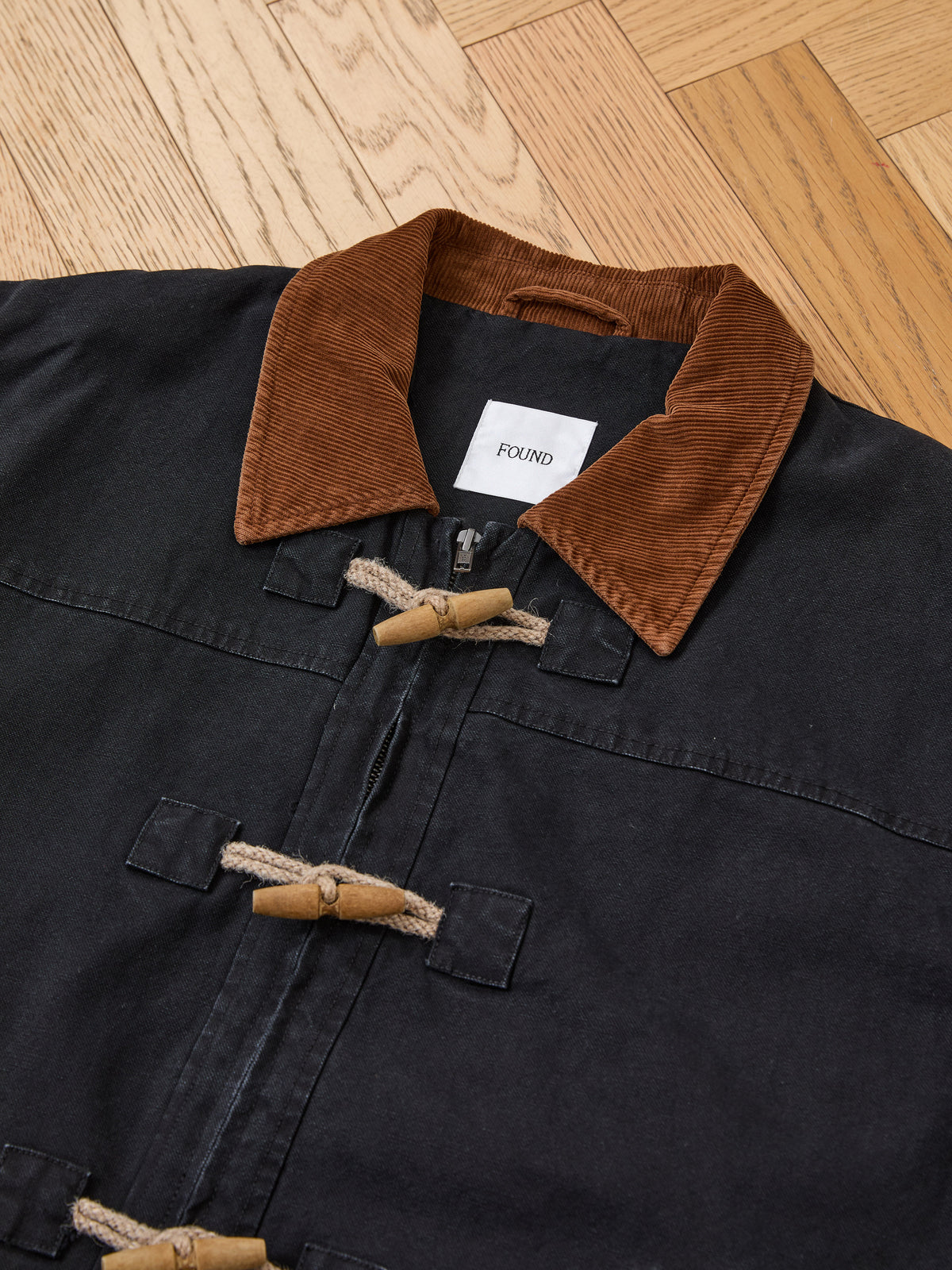 The Found Toggle Work Jacket features a black design with a brown corduroy collar, fireman-style toggle closures, and a distinctive white “FOUND” label—shown on a wooden floor, this vintage-inspired piece offers timeless workwear appeal.