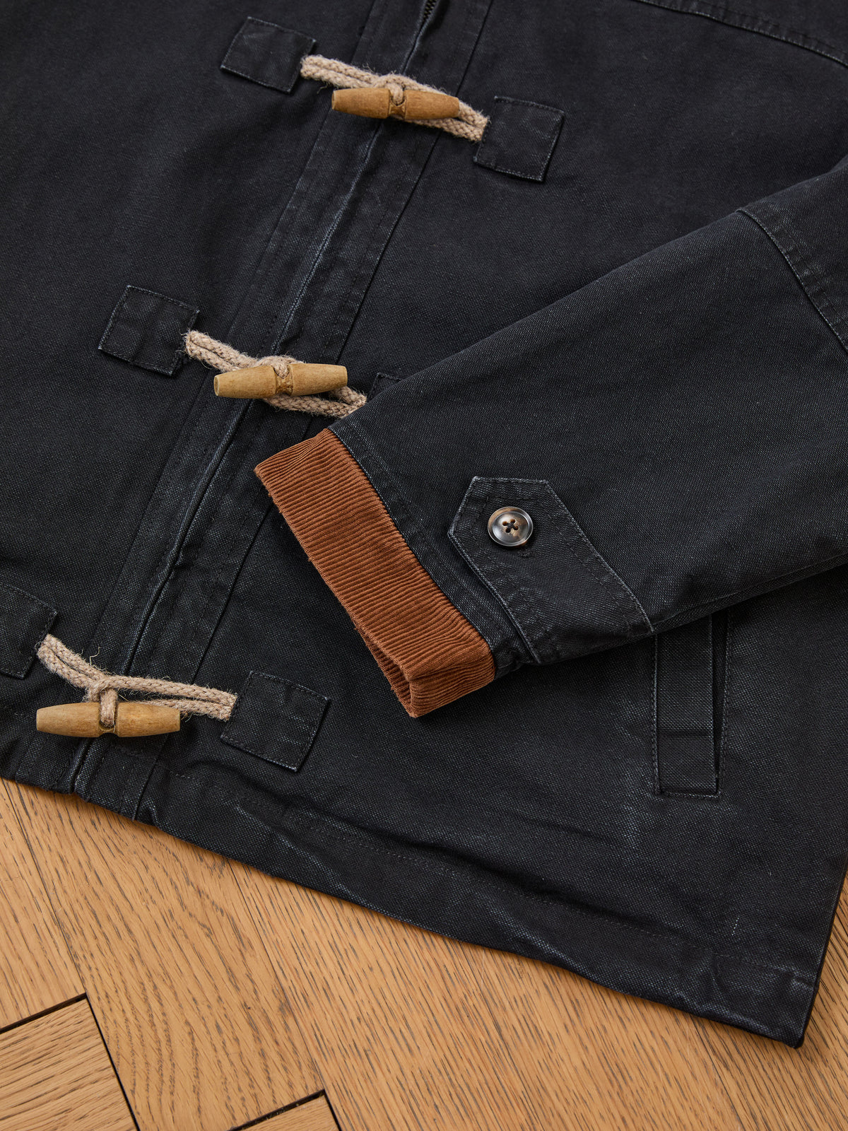 Close-up of the Found Toggle Work Jacket, a vintage-inspired unisex piece in a relaxed fit, featuring fireman-style wooden toggle closures, brown corduroy lining at the cuffs, and buttoned sleeves, laid flat on a wooden floor.