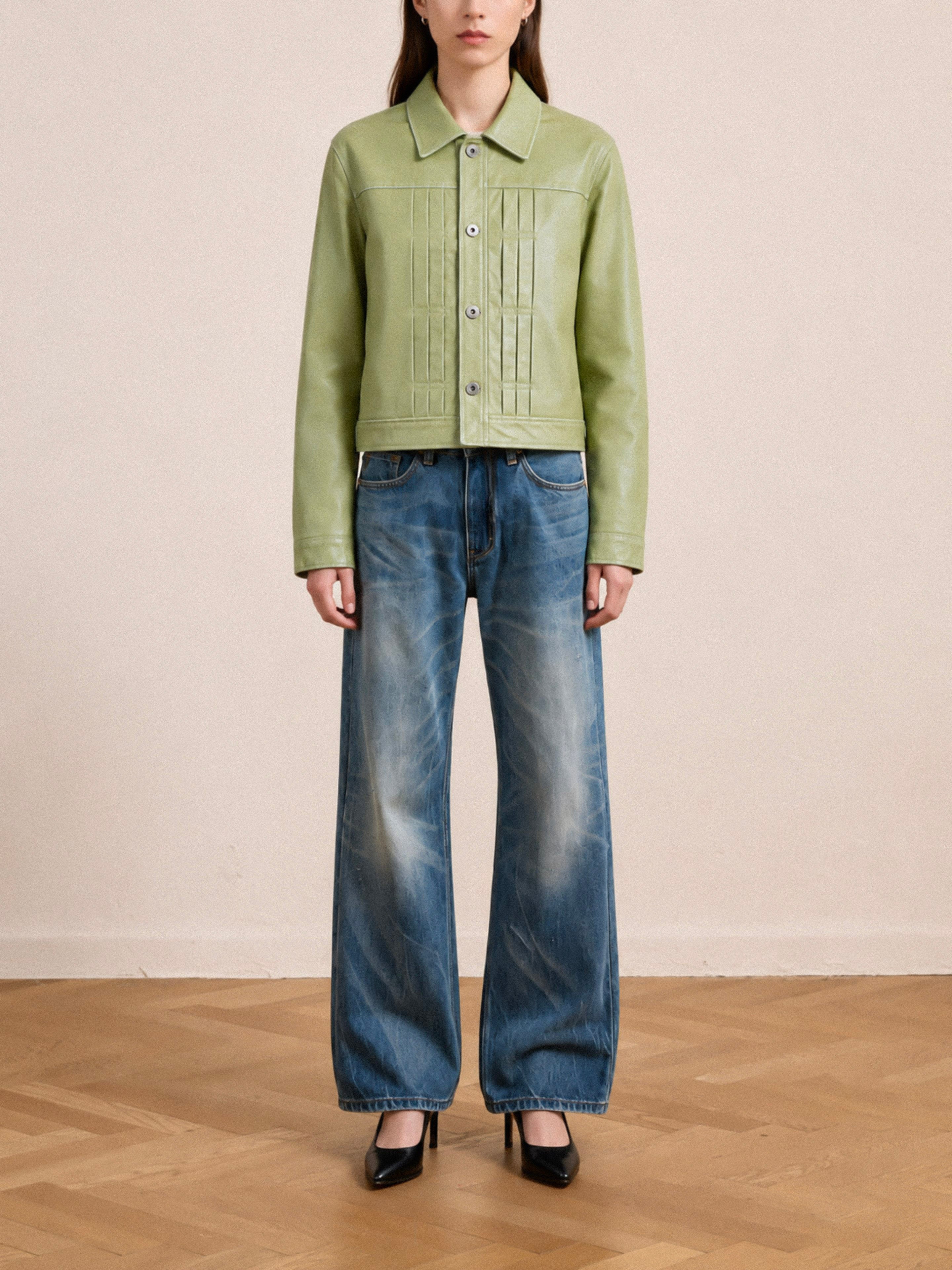A person stands indoors on a wooden floor against a plain beige background, wearing the FOUND Trucker Leather Jacket in washed green, styled with wide-leg blue jeans and black high-heeled shoes.