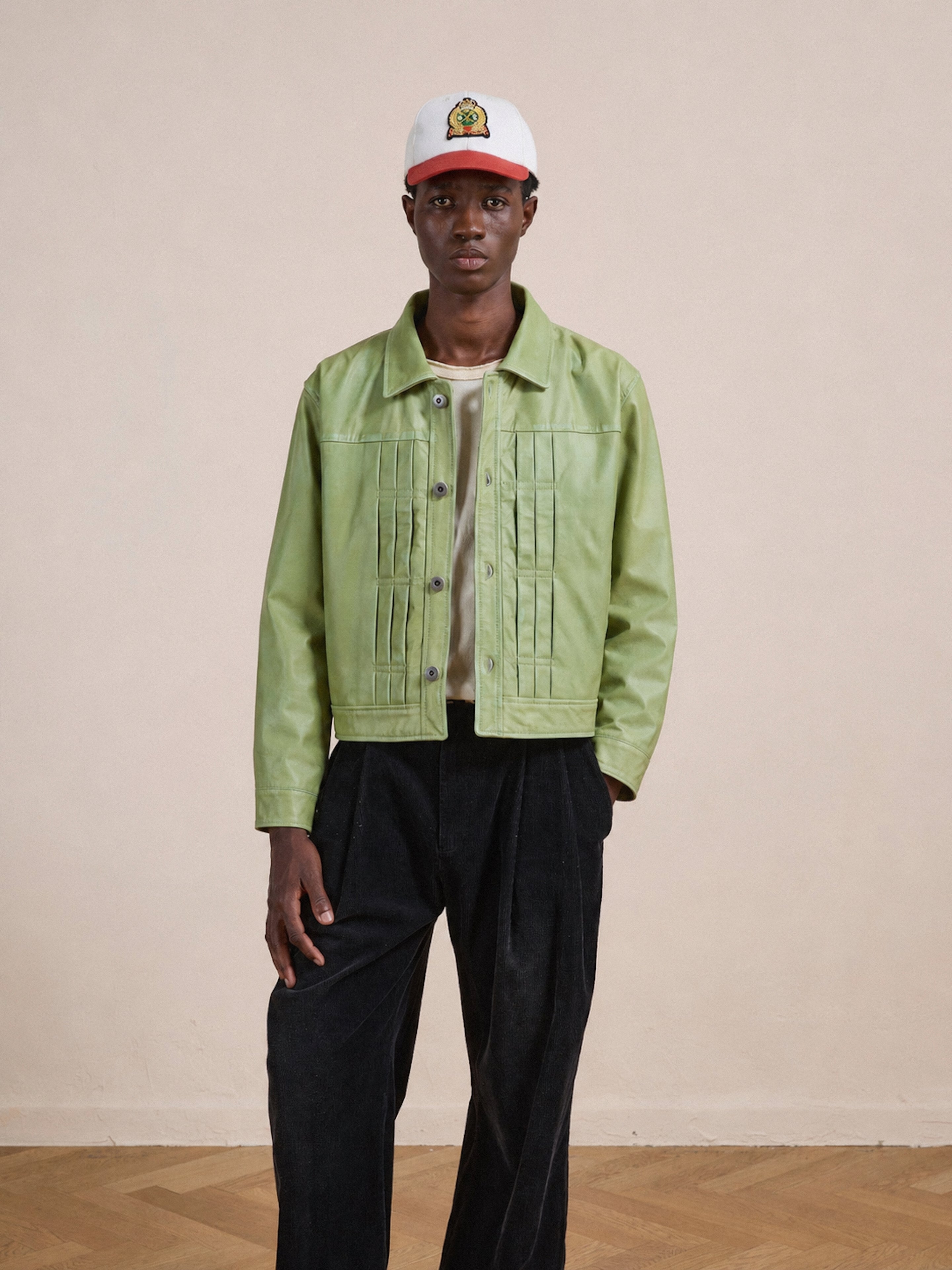 A person stands indoors facing the camera against a plain beige background, wearing the FOUND Trucker Leather Jacket in light green with a unisex relaxed fit, styled with black pants and a white baseball cap featuring a small colorful logo.