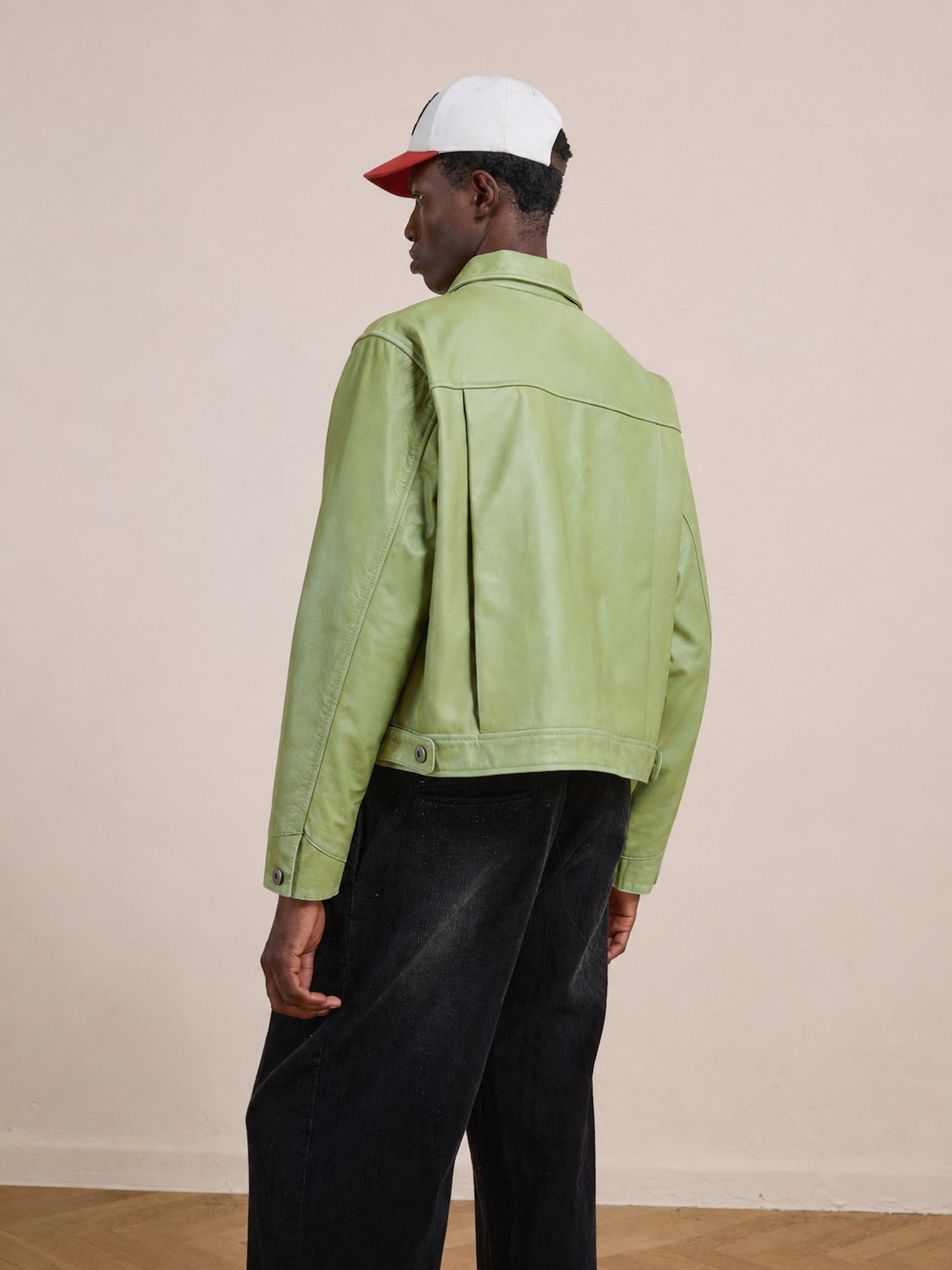 A person stands with their back to the camera against a plain background, wearing the FOUND Trucker Leather Jacket in light green, styled in a unisex relaxed fit, paired with black pants and a white and red baseball cap.