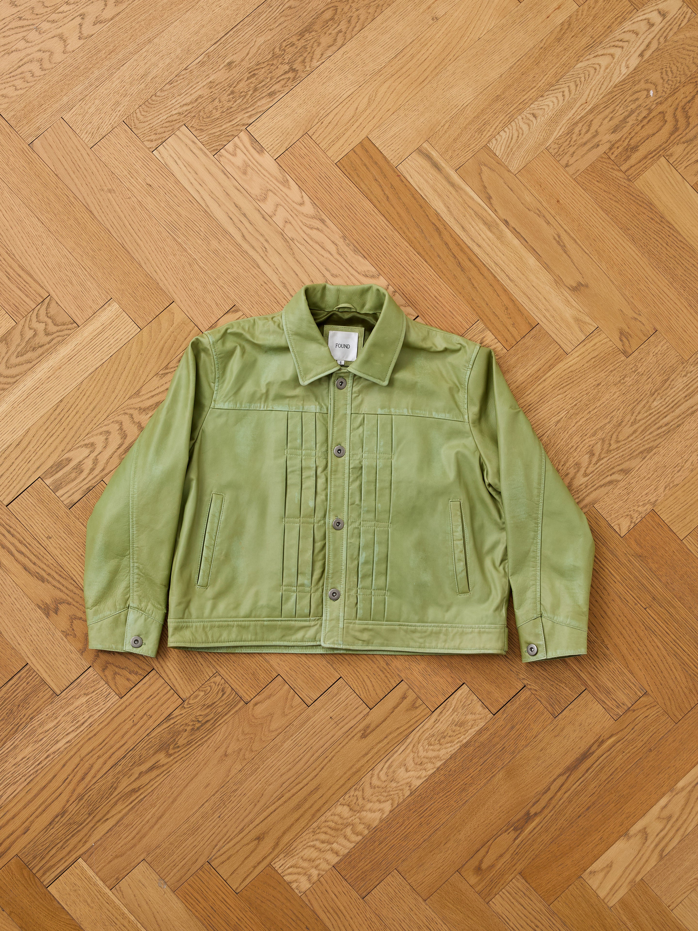 The FOUND Trucker Leather Jacket is a unisex, washed green piece with pleated details, button-up front, and relaxed fit, shown laid flat on a wooden parquet floor.