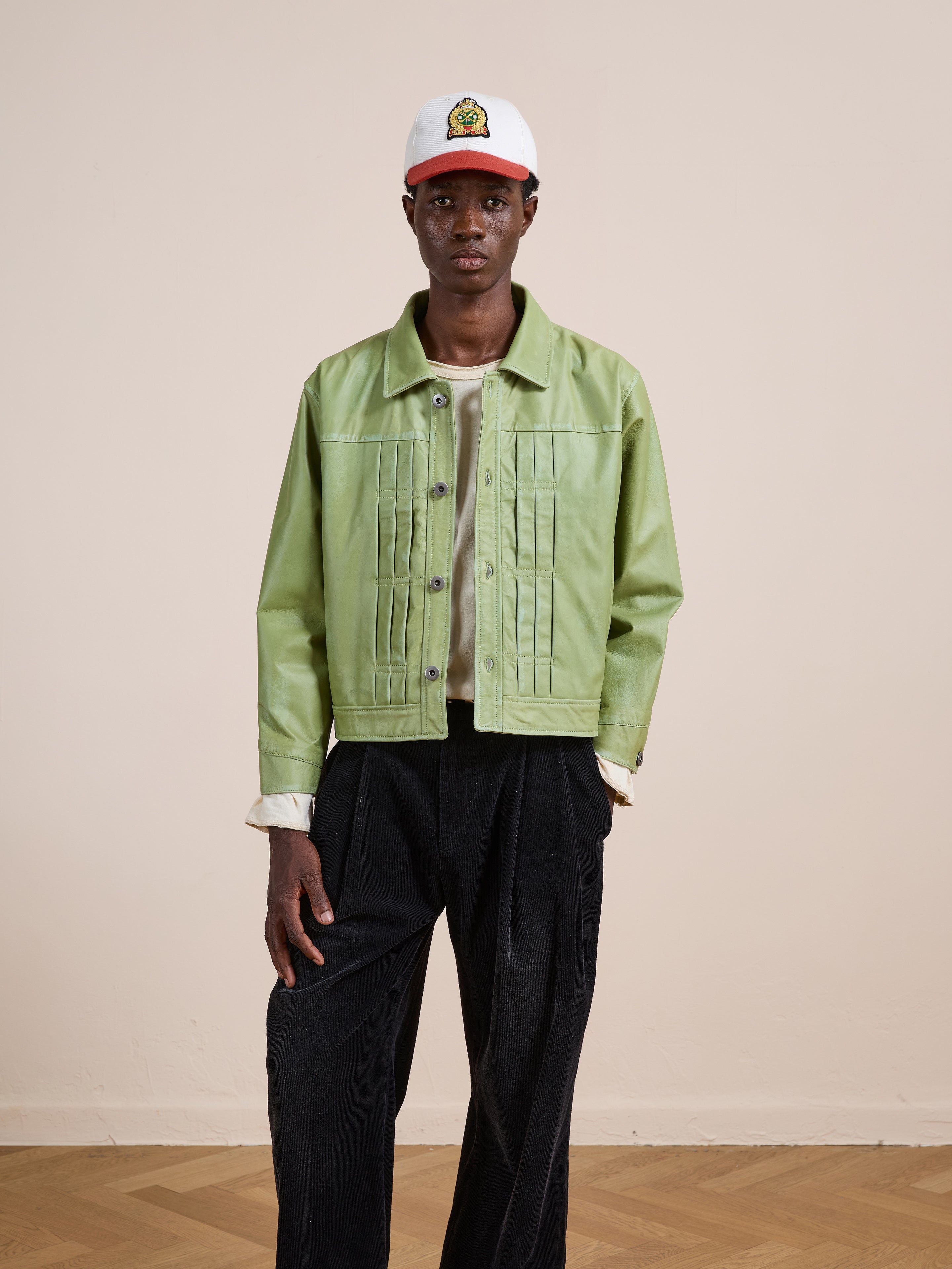 A person stands indoors against a beige wall and parquet floor, wearing FOUND's Trucker Leather Jacket in washed green, styled with black pants, a white shirt, and a white patch cap.