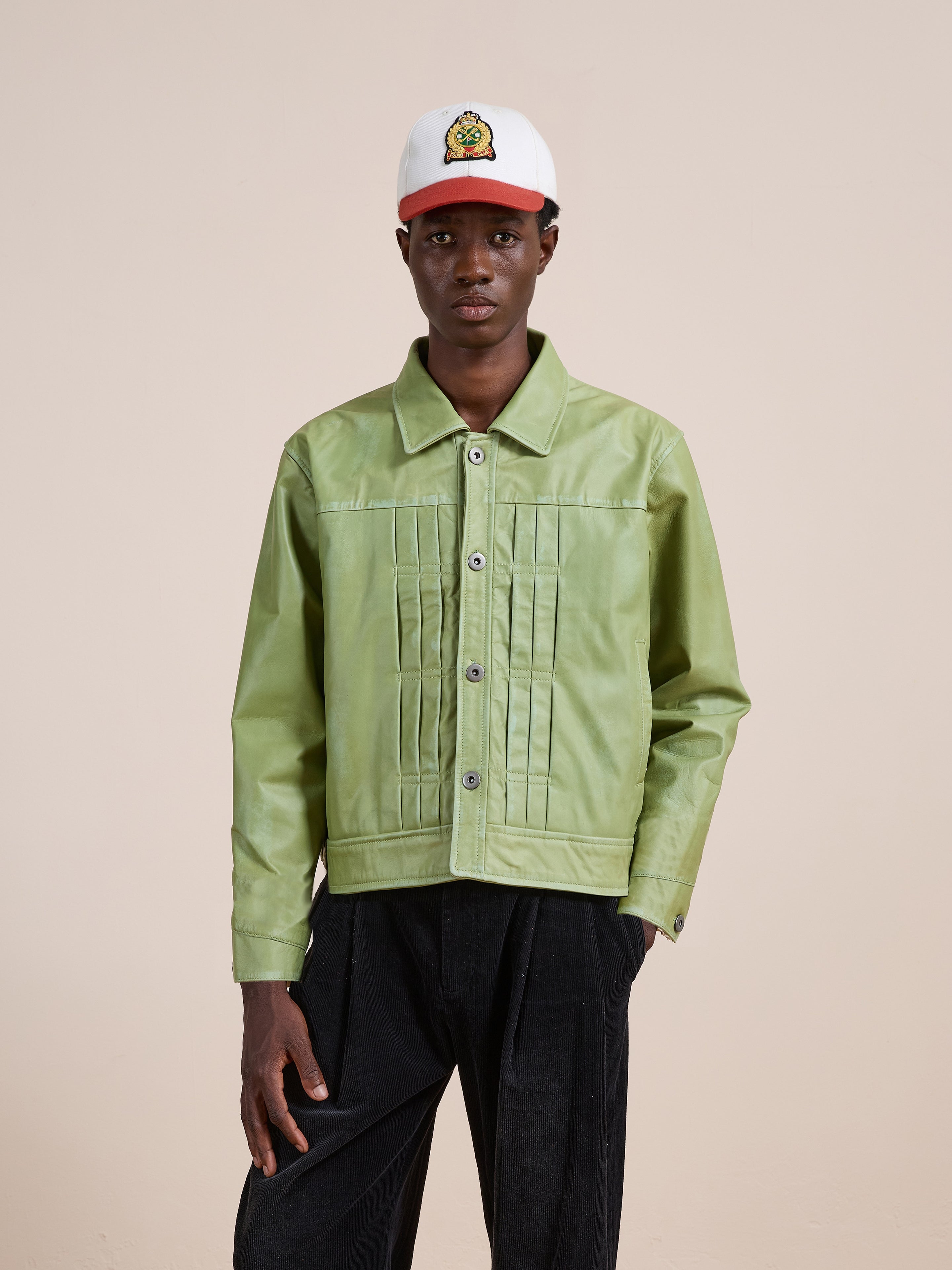 A person wearing the FOUND Trucker Leather Jacket in light green, featuring a unisex relaxed fit, pairs it with black pants and a white baseball cap adorned with a colorful patch while standing against a plain beige background.