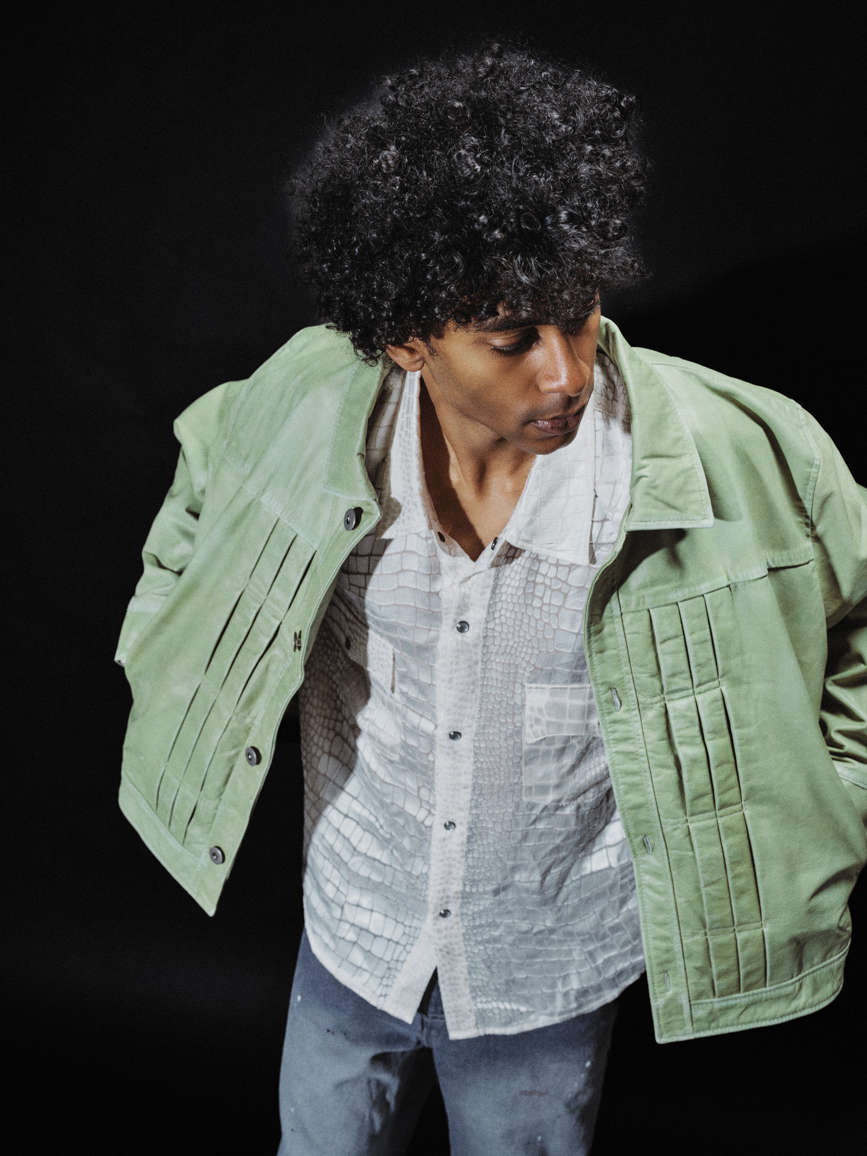 A person with curly hair wears the FOUND Trucker Leather Jacket in washed green over a patterned white shirt and blue jeans, standing against a dark background.