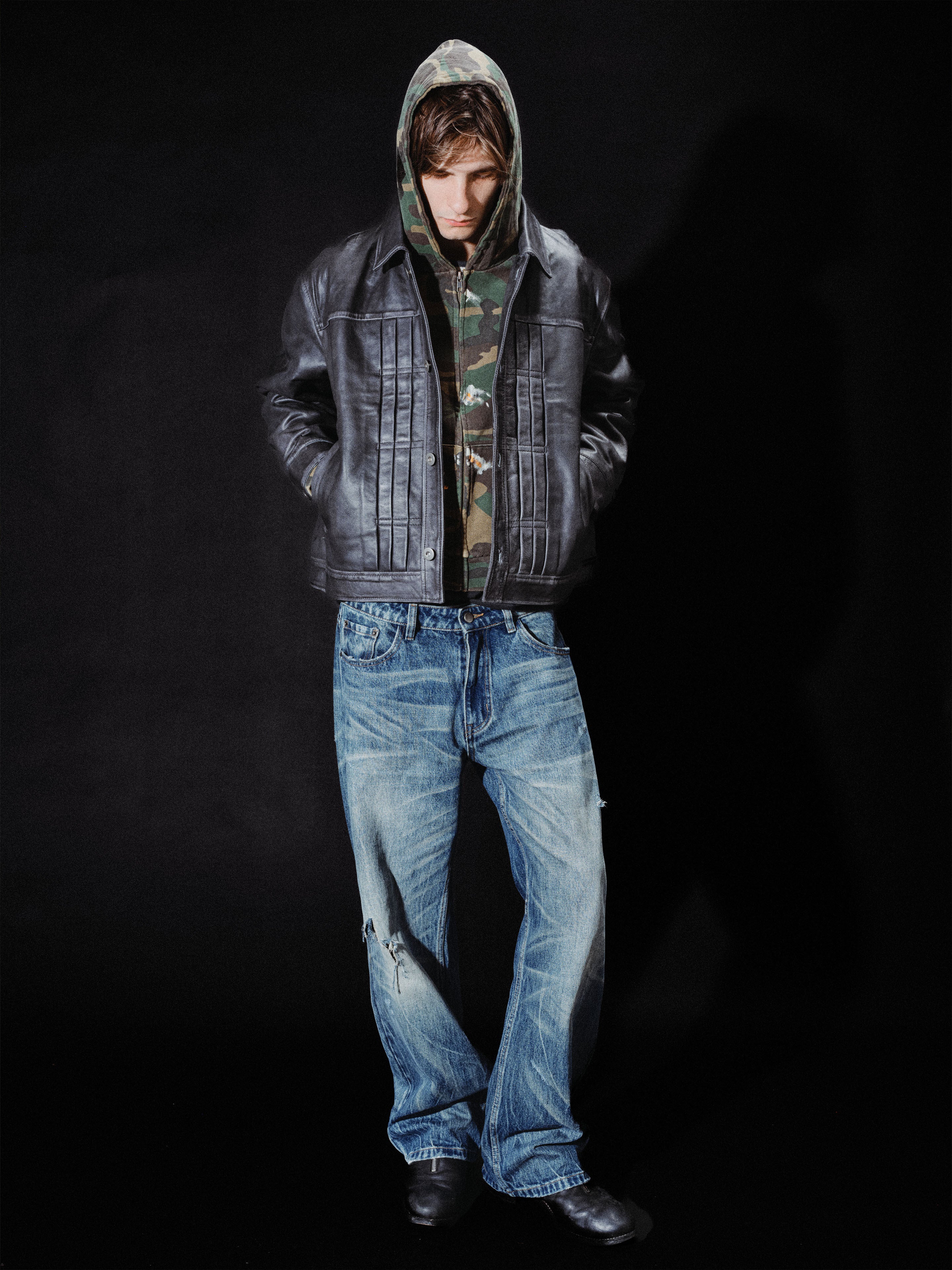 A person wearing the FOUND Trucker Leather Jacket, a camouflage hoodie, blue jeans, and black shoes stands against a black background with hands in their pockets.