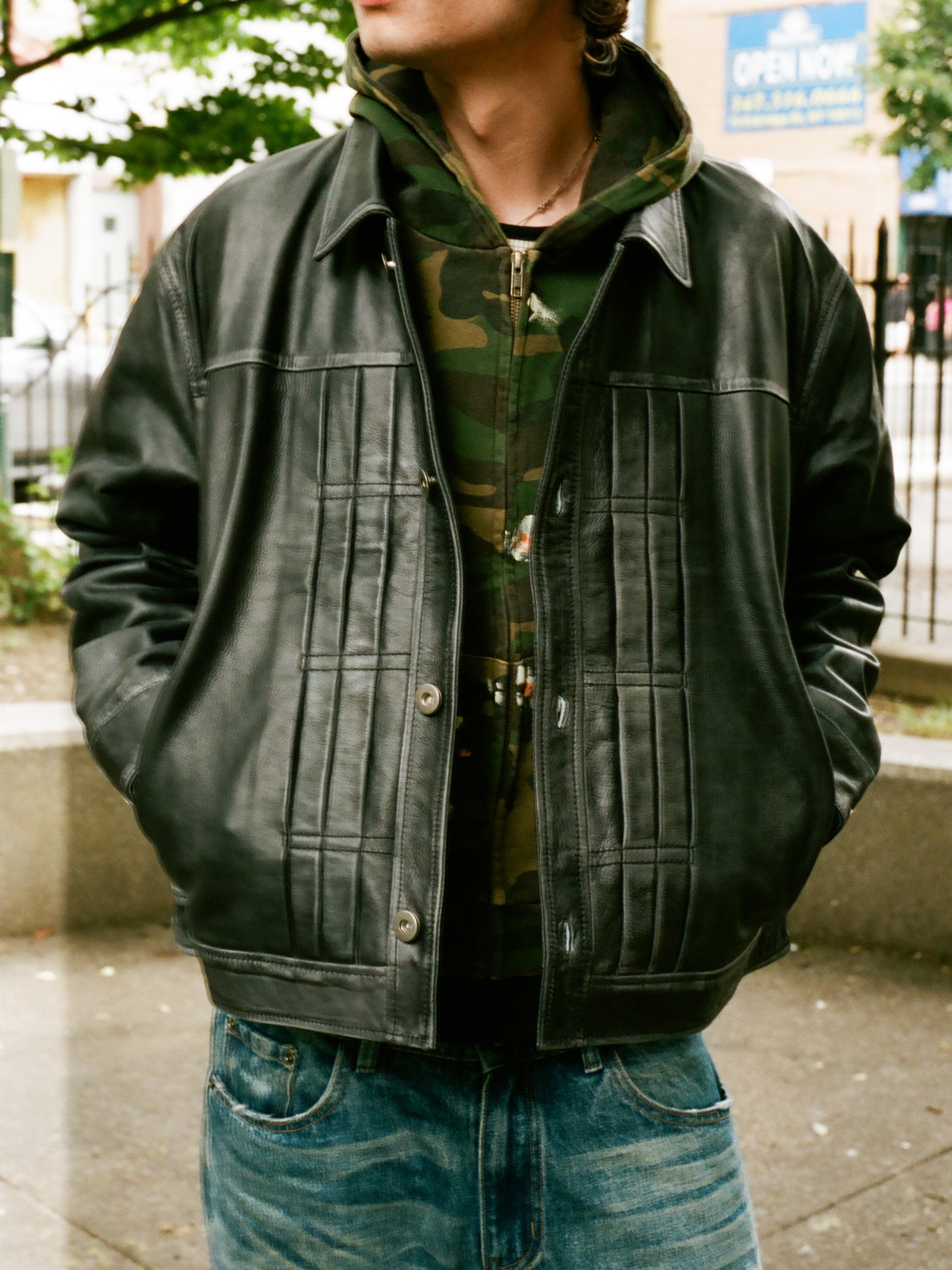 Wearing FOUND’s Trucker Leather Jacket over a camo hoodie and blue jeans, a person stands outdoors with hands in their pockets.