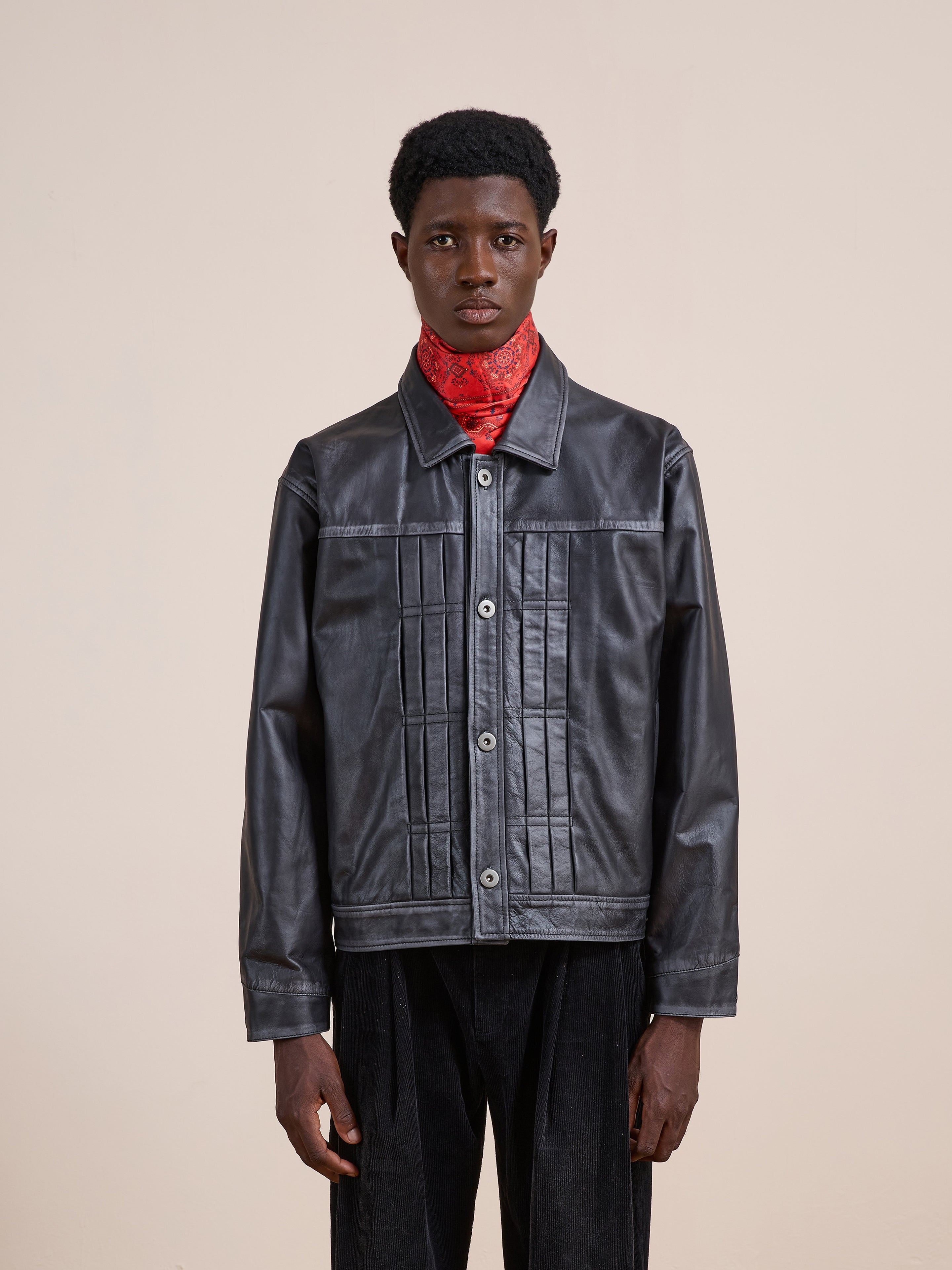 A person stands facing forward, wearing the FOUND Trucker Leather Jacket—black leather with vertical stitching—and a red patterned scarf, against a plain beige background.