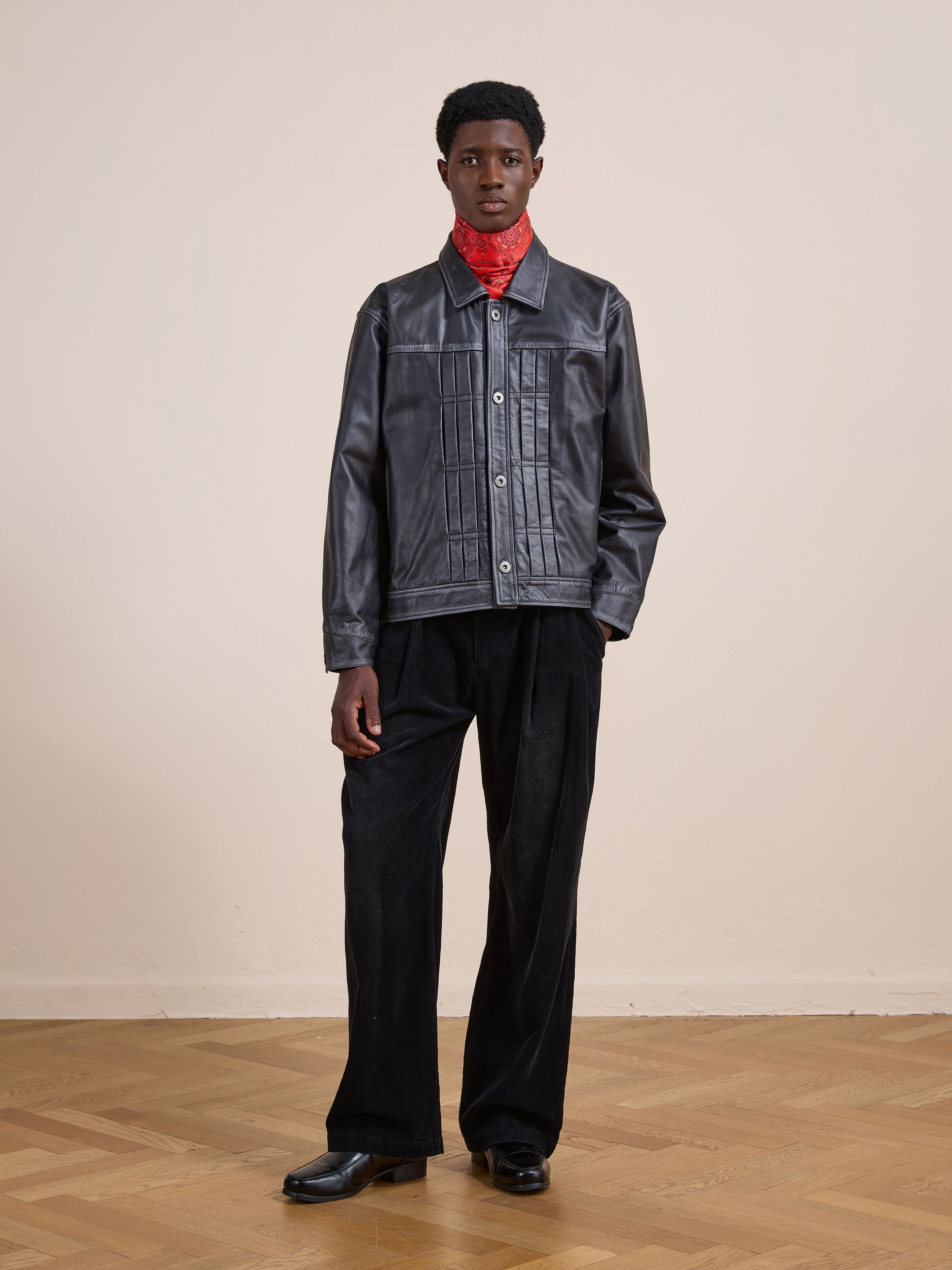 Indoors on a wooden floor, a person wears the FOUND Trucker Leather Jacket with a relaxed fit, styled with black pants, black shoes, and a red bandana around the neck, standing before a plain beige wall.