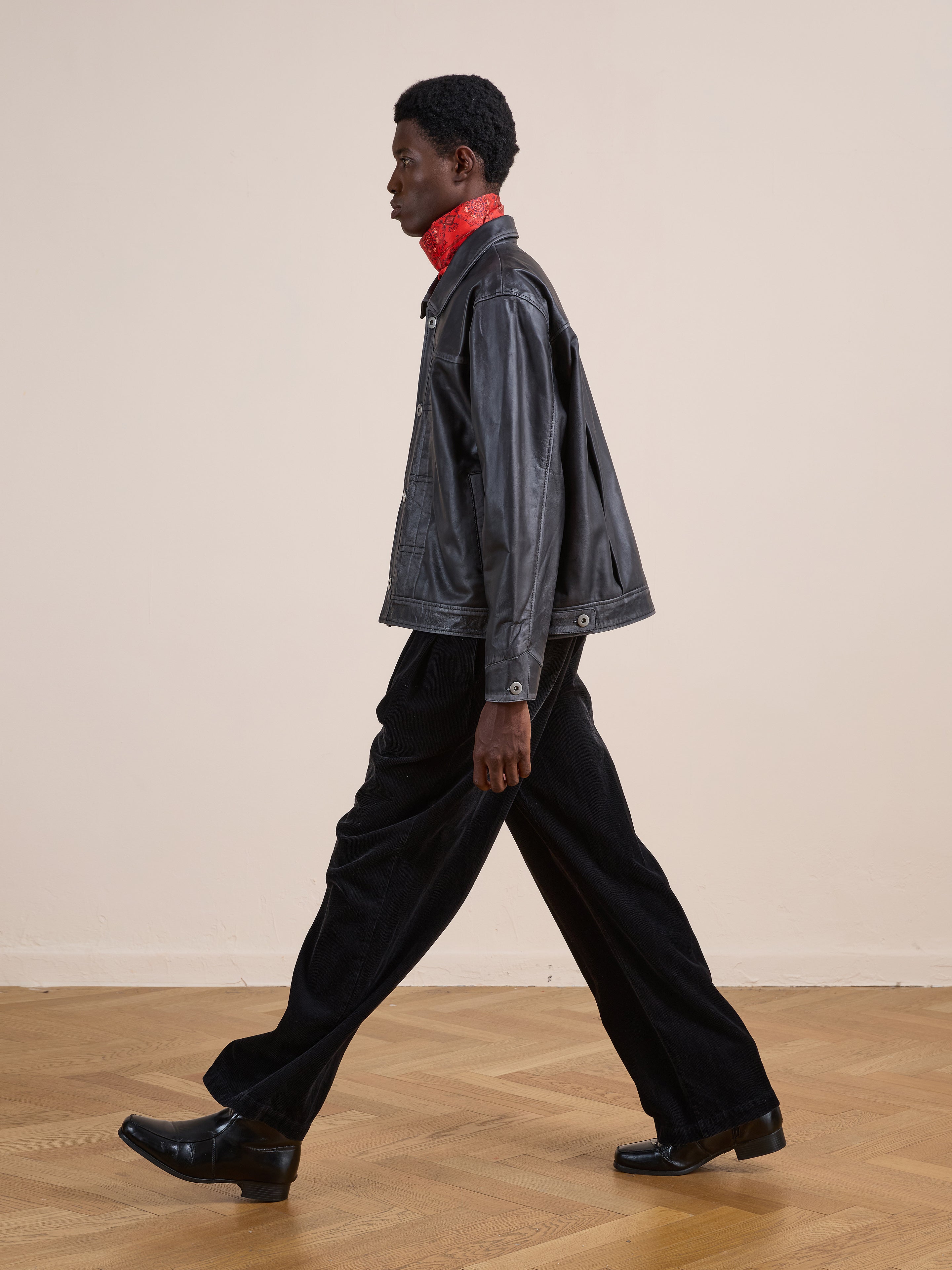 A person indoors wearing the FOUND Trucker Leather Jacket, styled with black pants, black shoes, and a red scarf, stands against a plain beige wall with wooden flooring.
