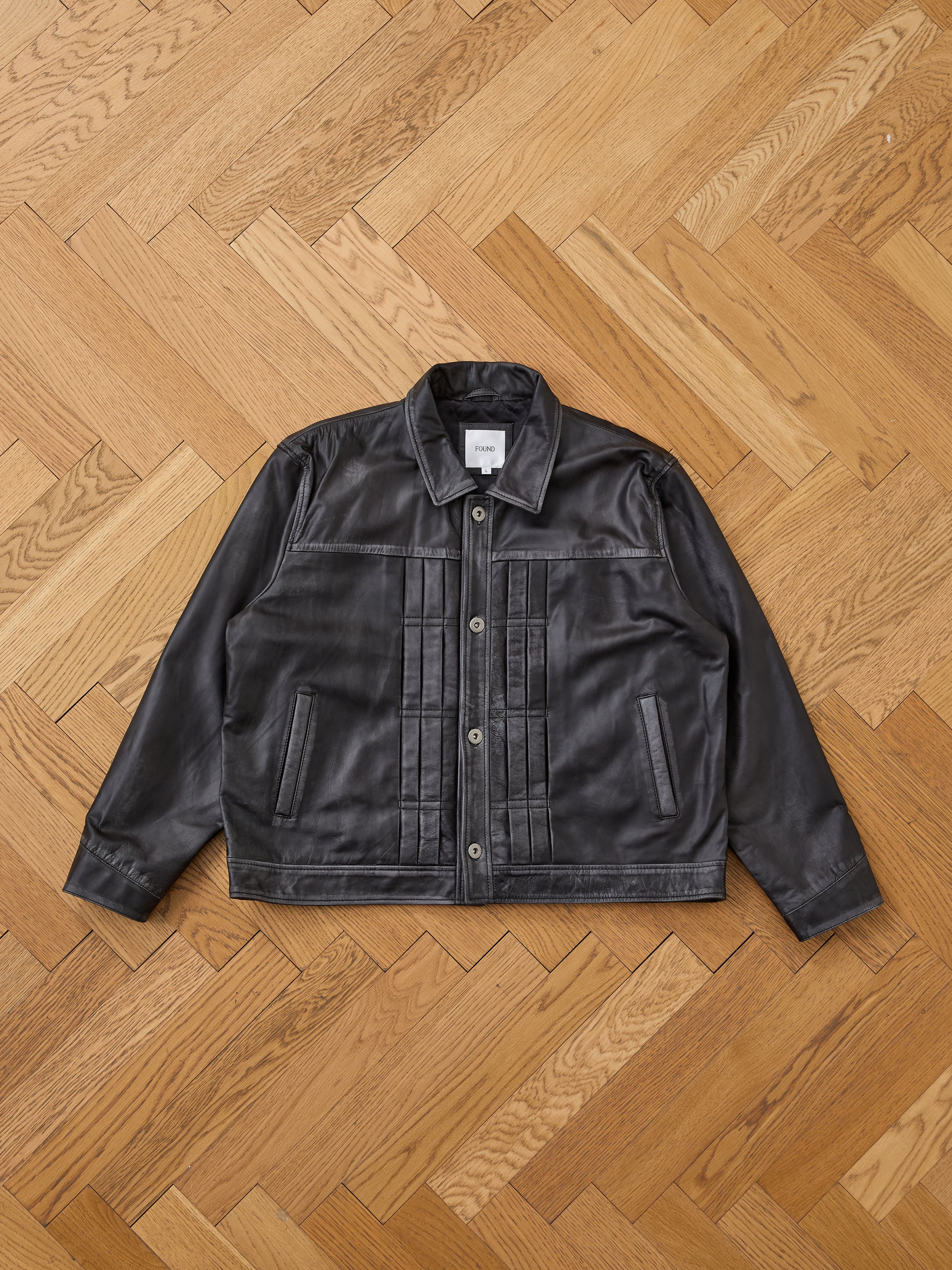The FOUND Trucker Leather Jacket, featuring a vintage black leather design with button closure and front pockets, is laid flat on a wooden herringbone-patterned floor.