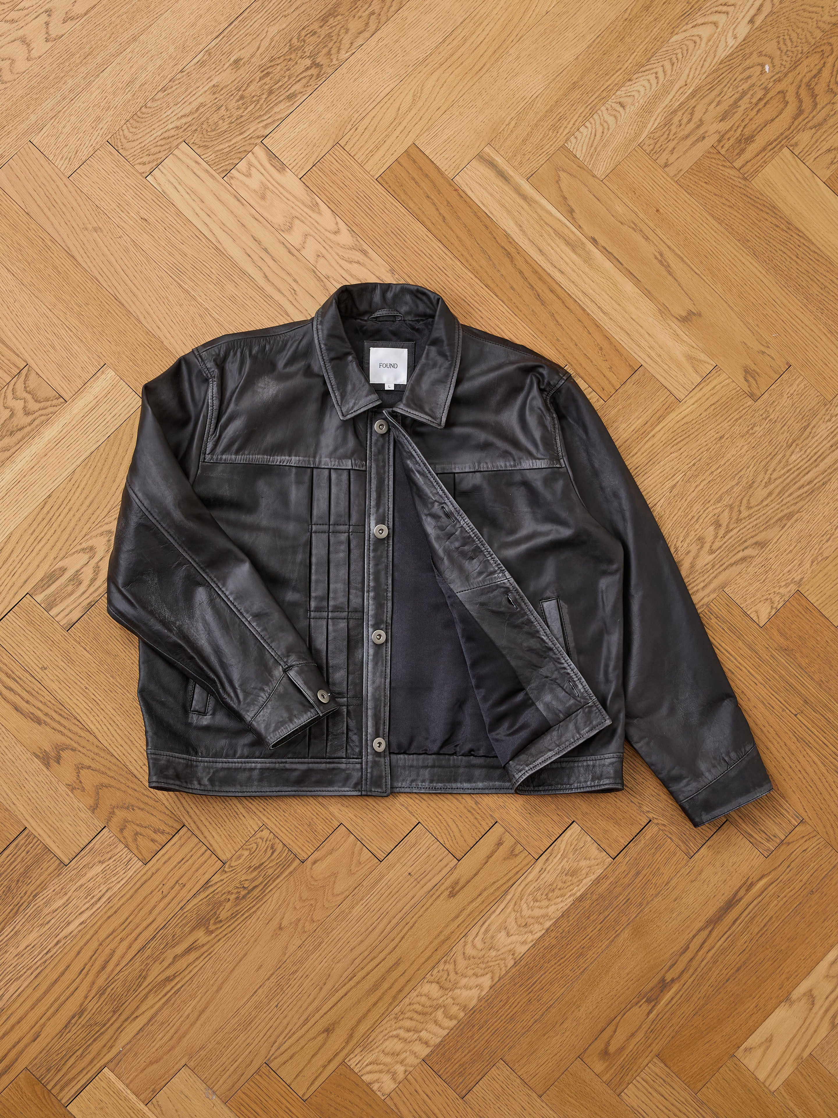 The FOUND Trucker Leather Jacket in black leather is displayed flat on a light wooden herringbone floor, with one side unbuttoned to reveal its inner lining.