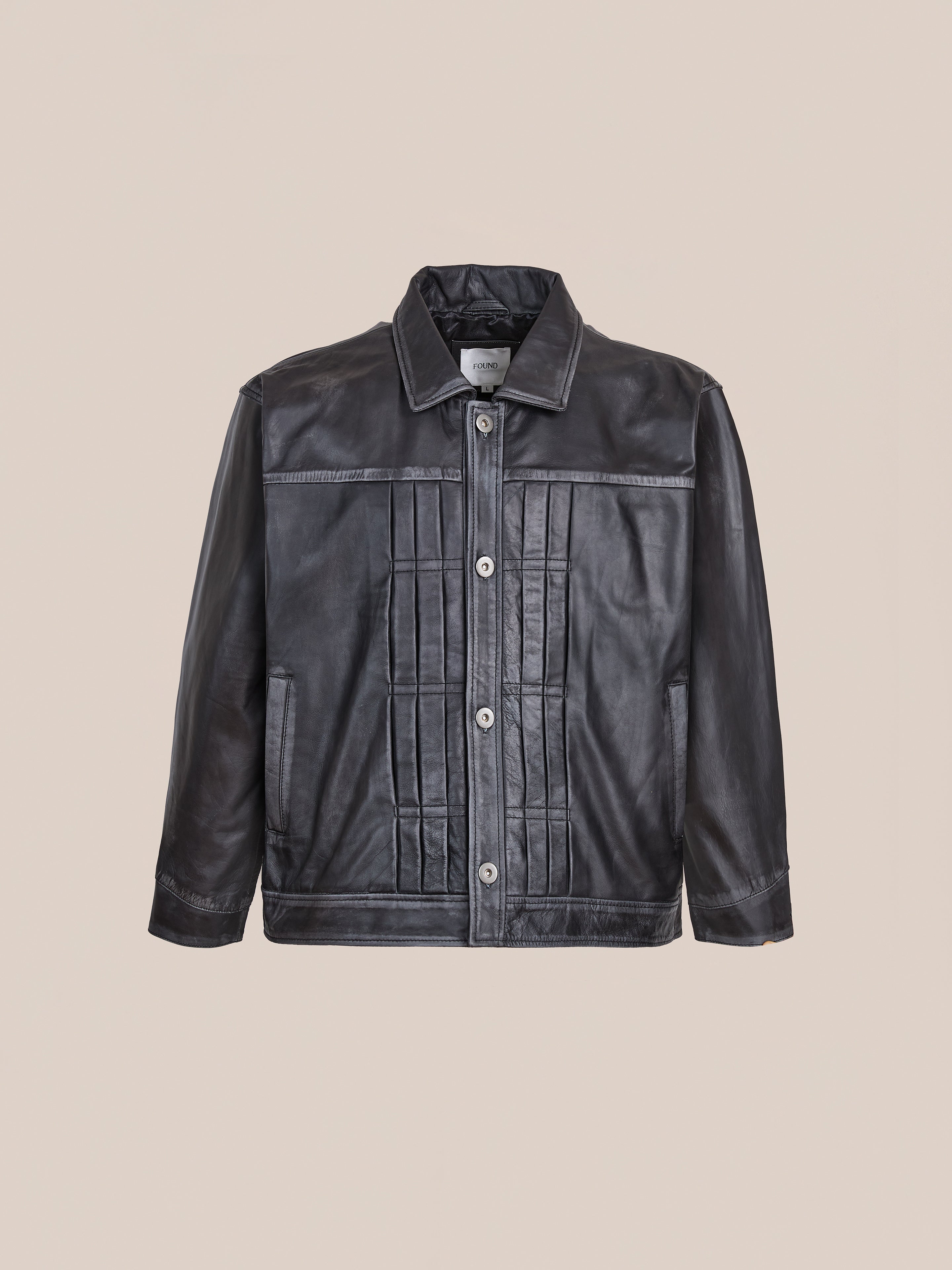 The FOUND Trucker Leather Jacket features a vintage black leather design with front button closure, pleated front panels, long sleeves, and a collar, shown against a plain beige background.