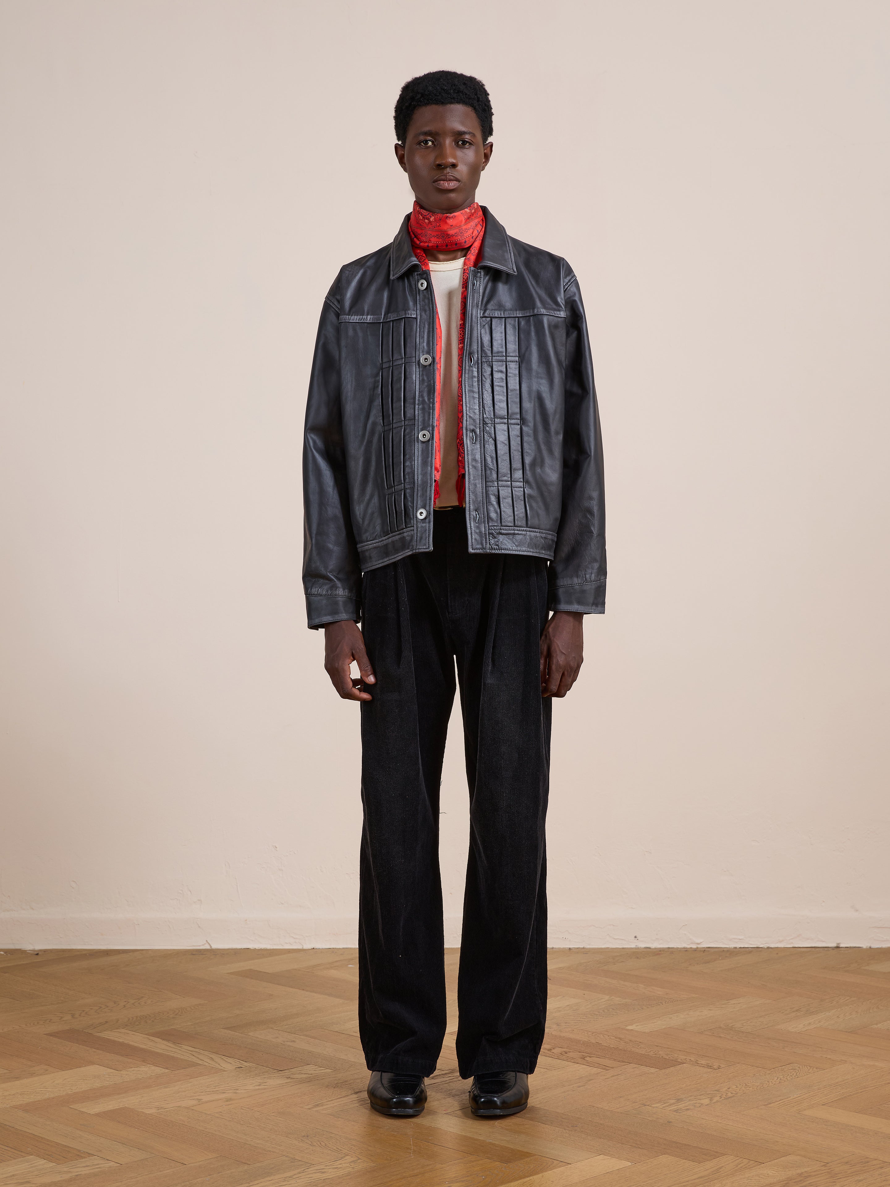 Person stands indoors against a plain light wall, wearing the FOUND Trucker Leather Jacket, paired with a red scarf, white shirt, black trousers, and black shoes, facing the camera.