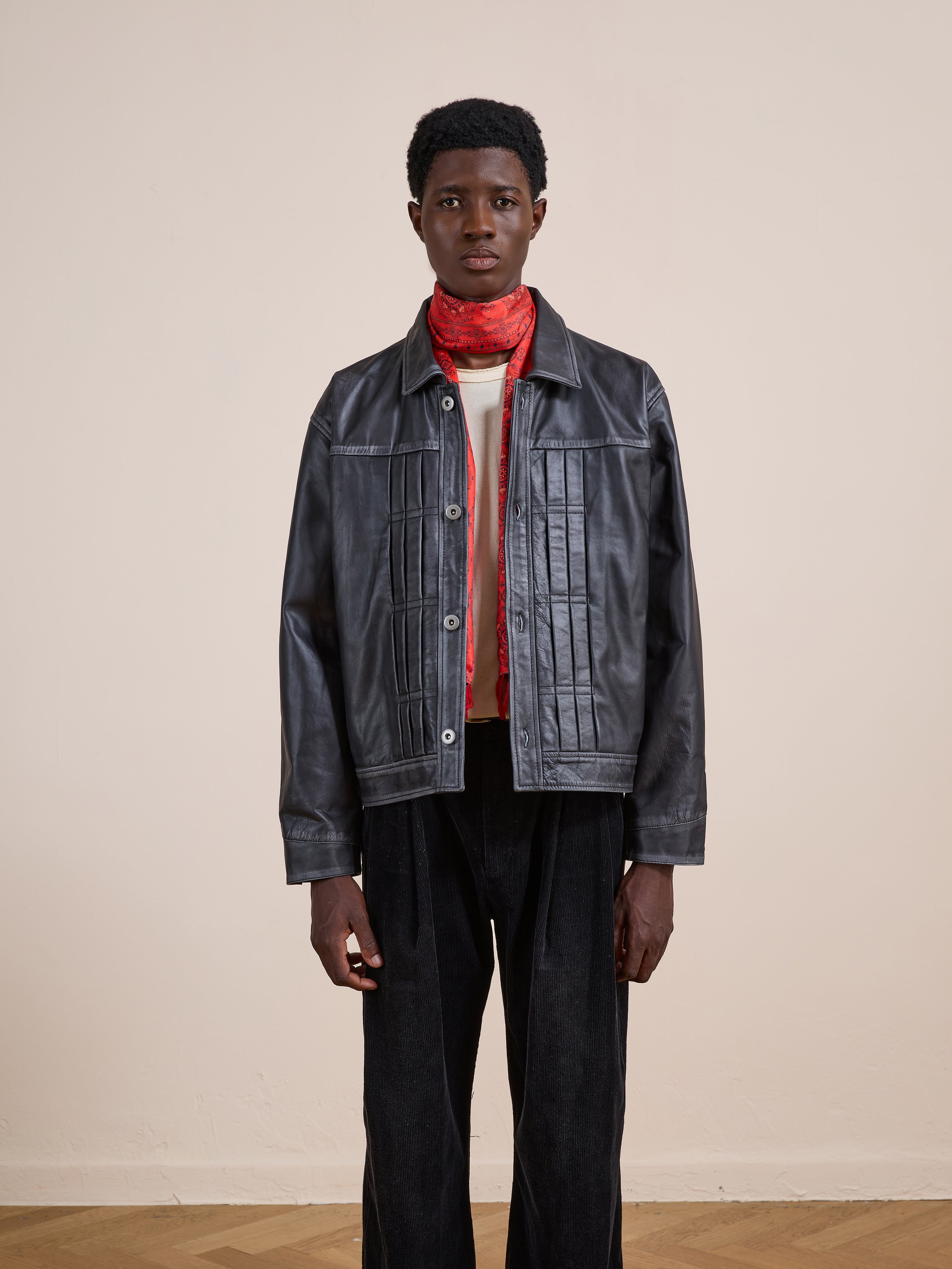 A person stands against a plain wall wearing FOUND's Trucker Leather Jacket, black pants, a white shirt, and a red scarf.